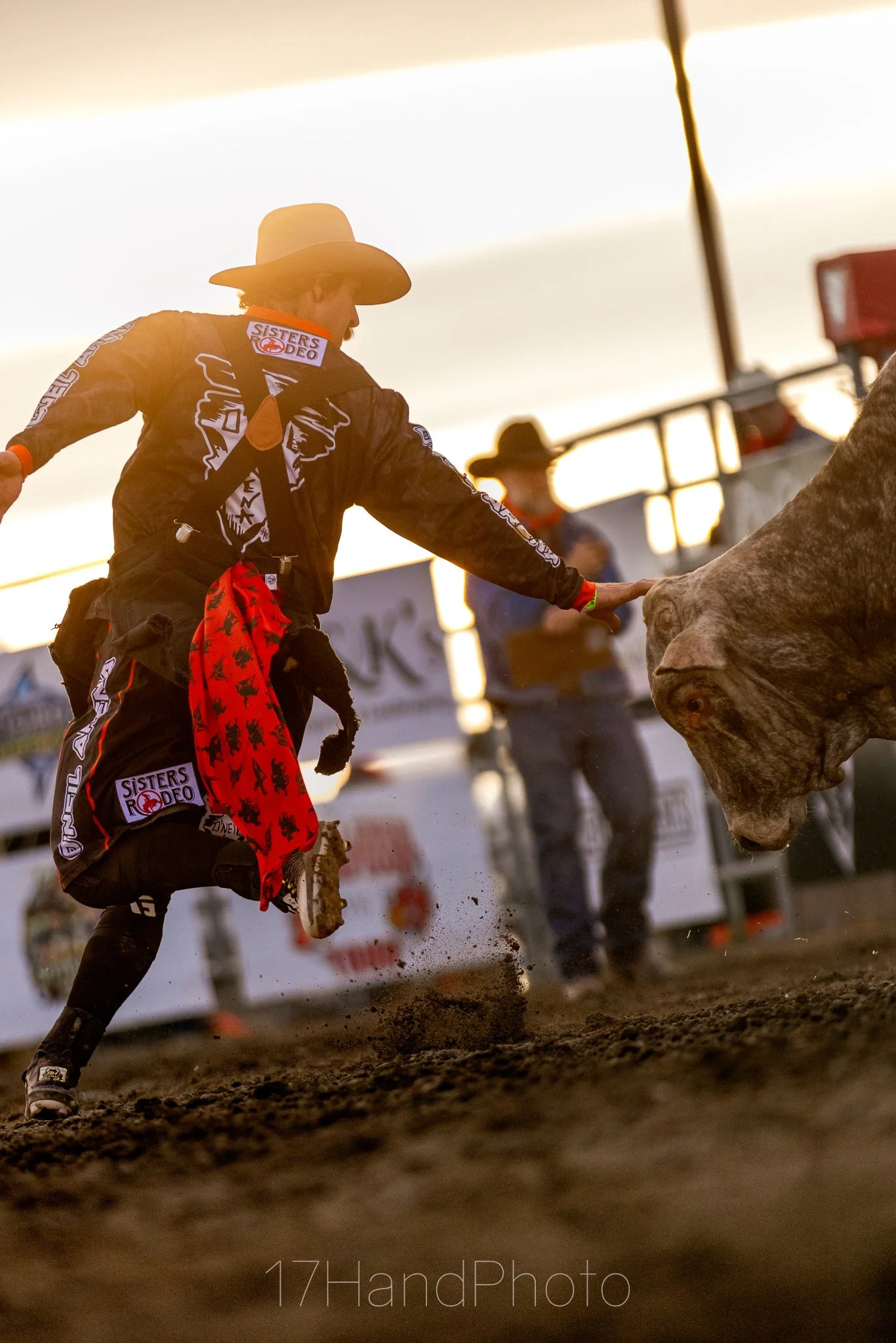 A rodeo cowboy in a black suit and wide-brimmed hat, touching a bull during a rodeo event at sunset.