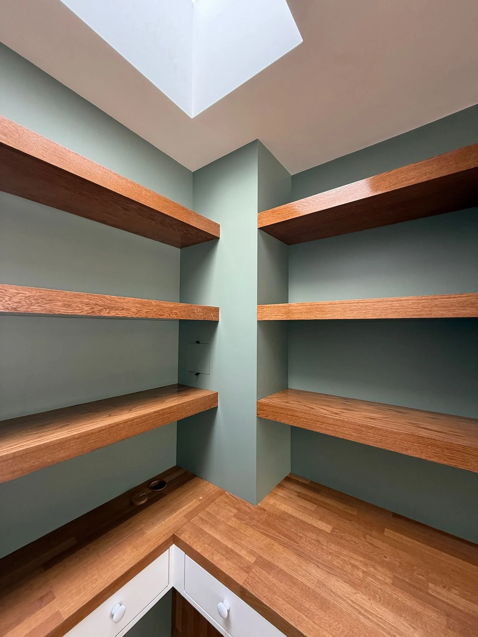 Bespoke thick wooden floating shelves mounted on sage green walls in an organized walk-in pantry.butcher block countertops illuminated by a modern square ceiling skylight