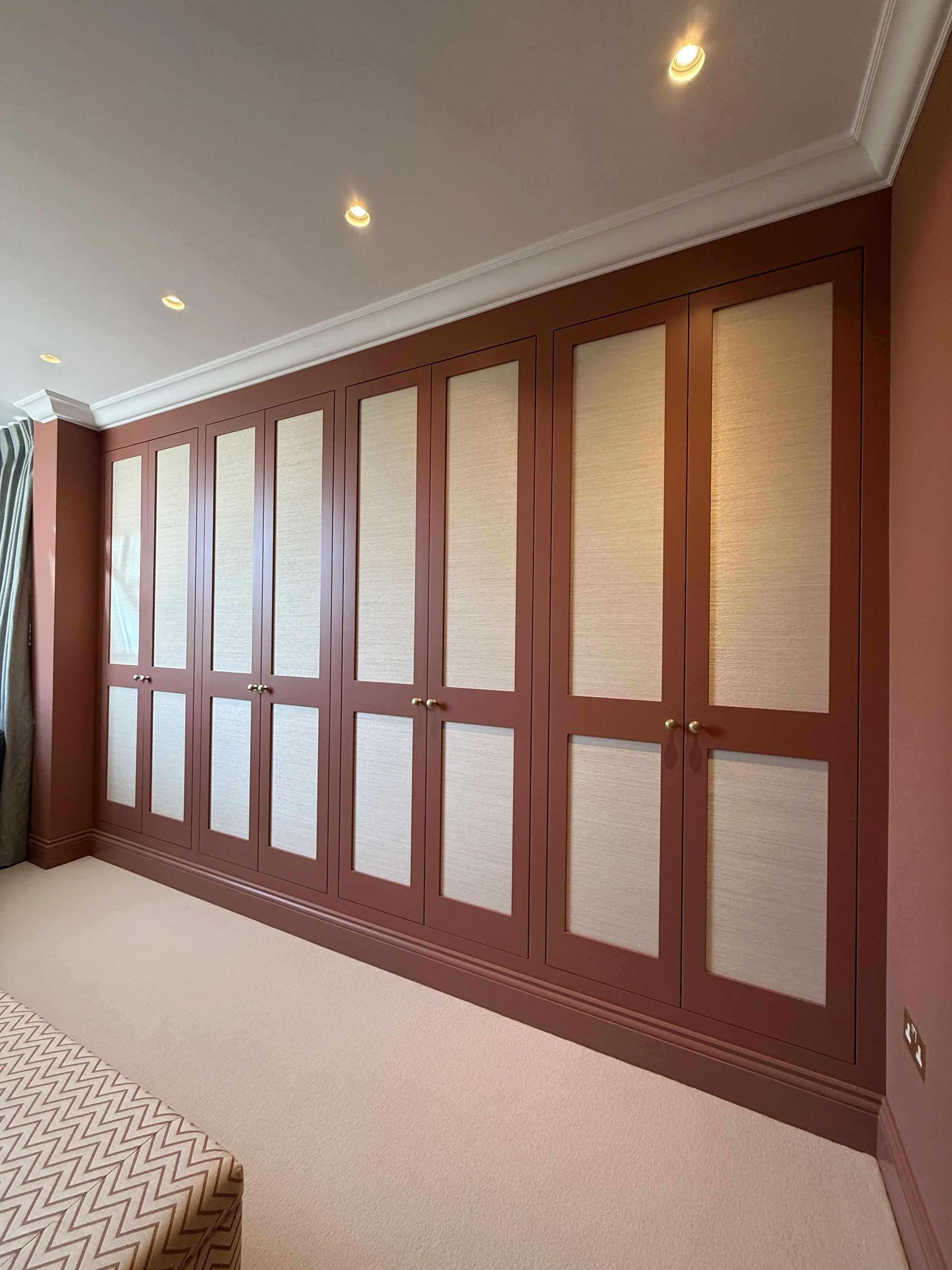 Modern bedroom featuring floor-to-ceiling clay-colored fitted wardrobes with woven fabric inserts and recessed lighting.