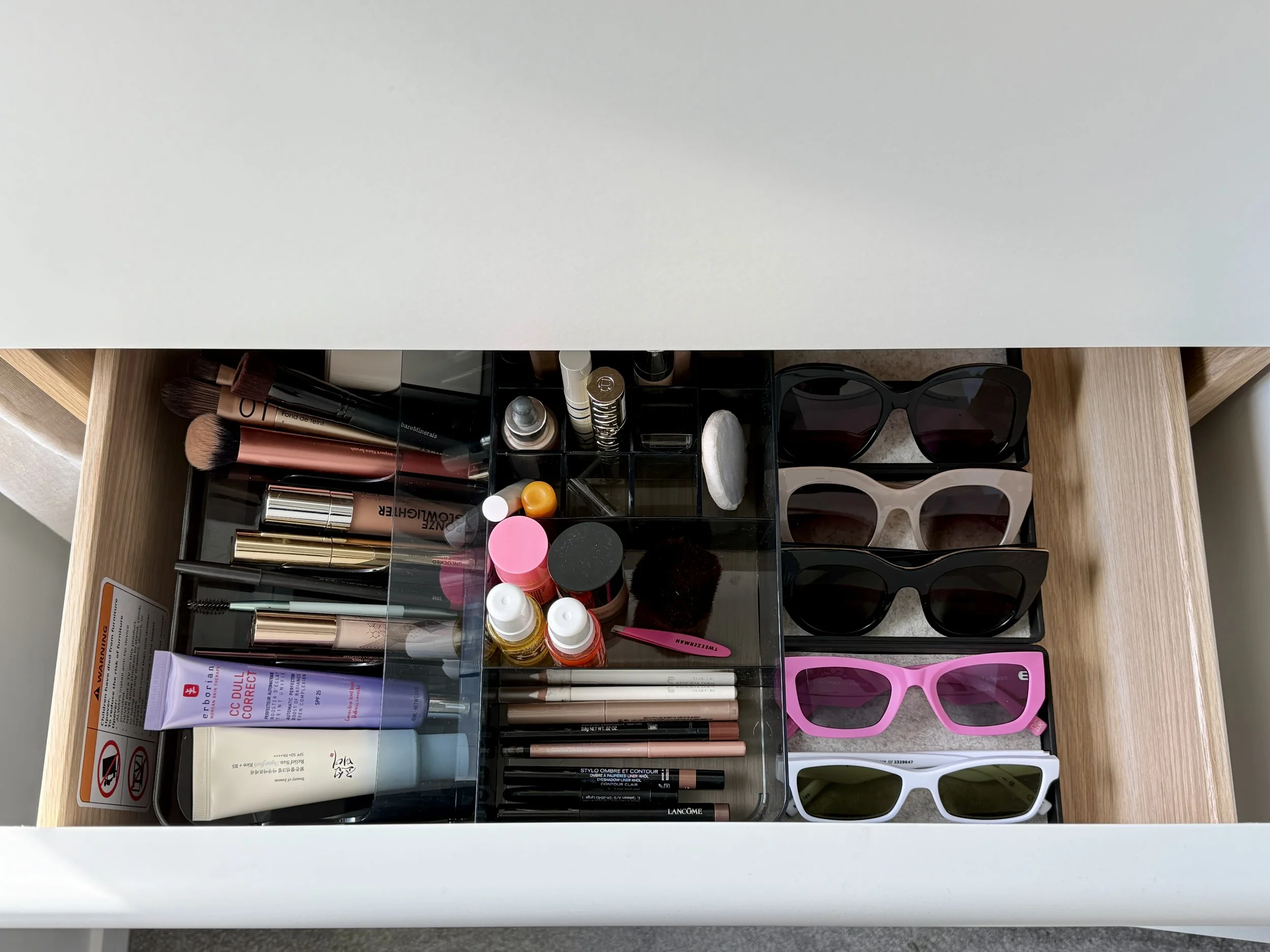 Vanity drawer organization using clear acrylic dividers to neatly separate makeup brushes, cosmetics, and sunglasses.