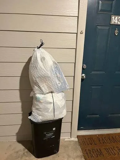 A black trash can placed outside a house with a beige siding wall and a blue door. Two large white plastic bags are stacked inside the trash can.