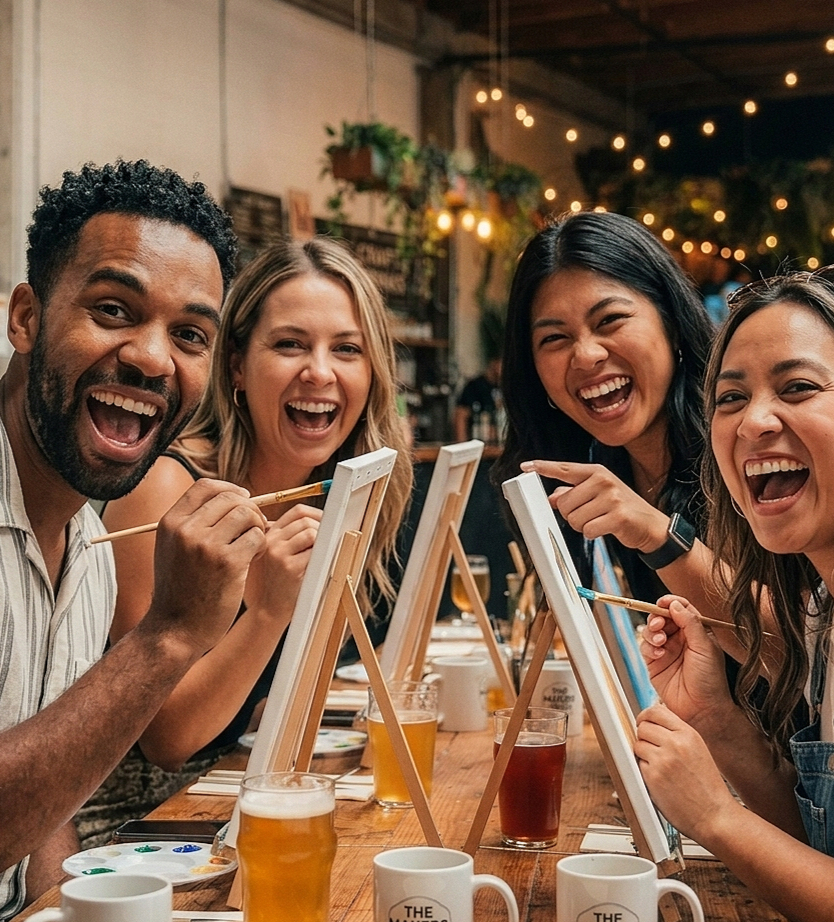 Group of four friends painting on canvases at a table in a cozy, warmly lit art studio or café.
