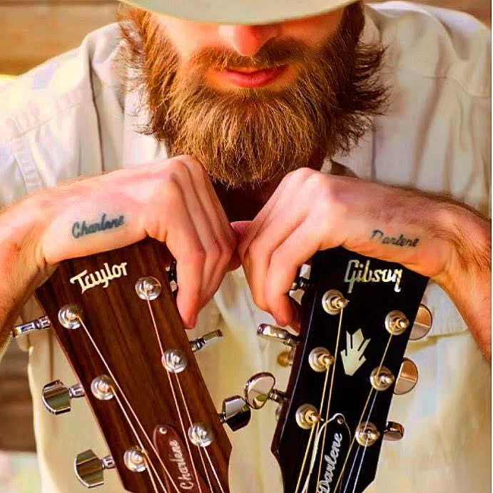 Close-up of man with a beard wearing a hat, holding two guitar heads with the brand names Taylor and Gibson.