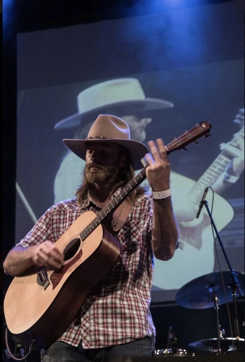 Musician playing an acoustic guitar on stage with a large screen behind him displaying a similar image of himself.