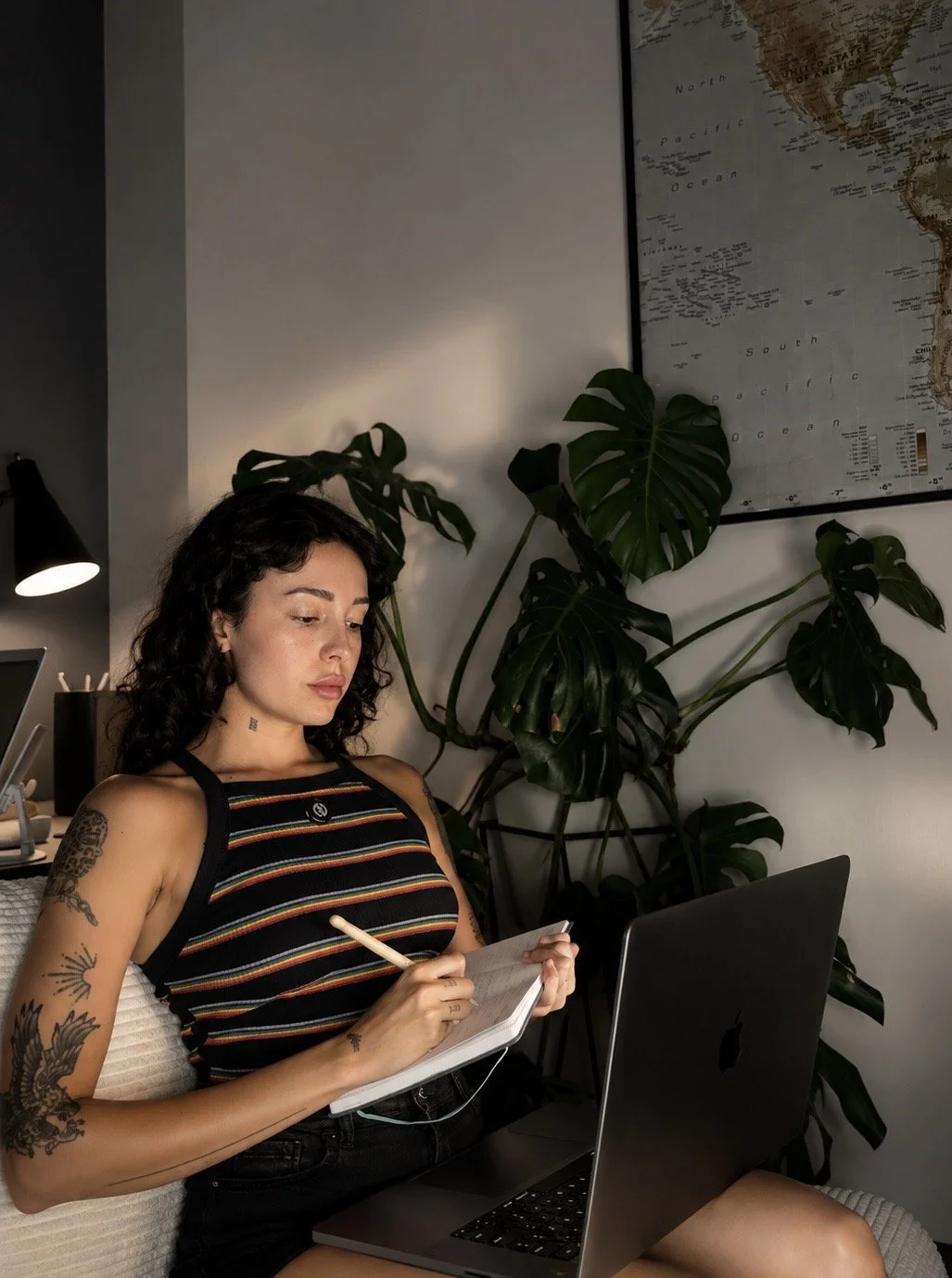 A woman with tattoos on her arms and shoulder, wearing a striped sleeveless top, sits at a desk with a laptop, writing in a notepad with a pen, in a room decorated with green houseplants and a world map on the wall.