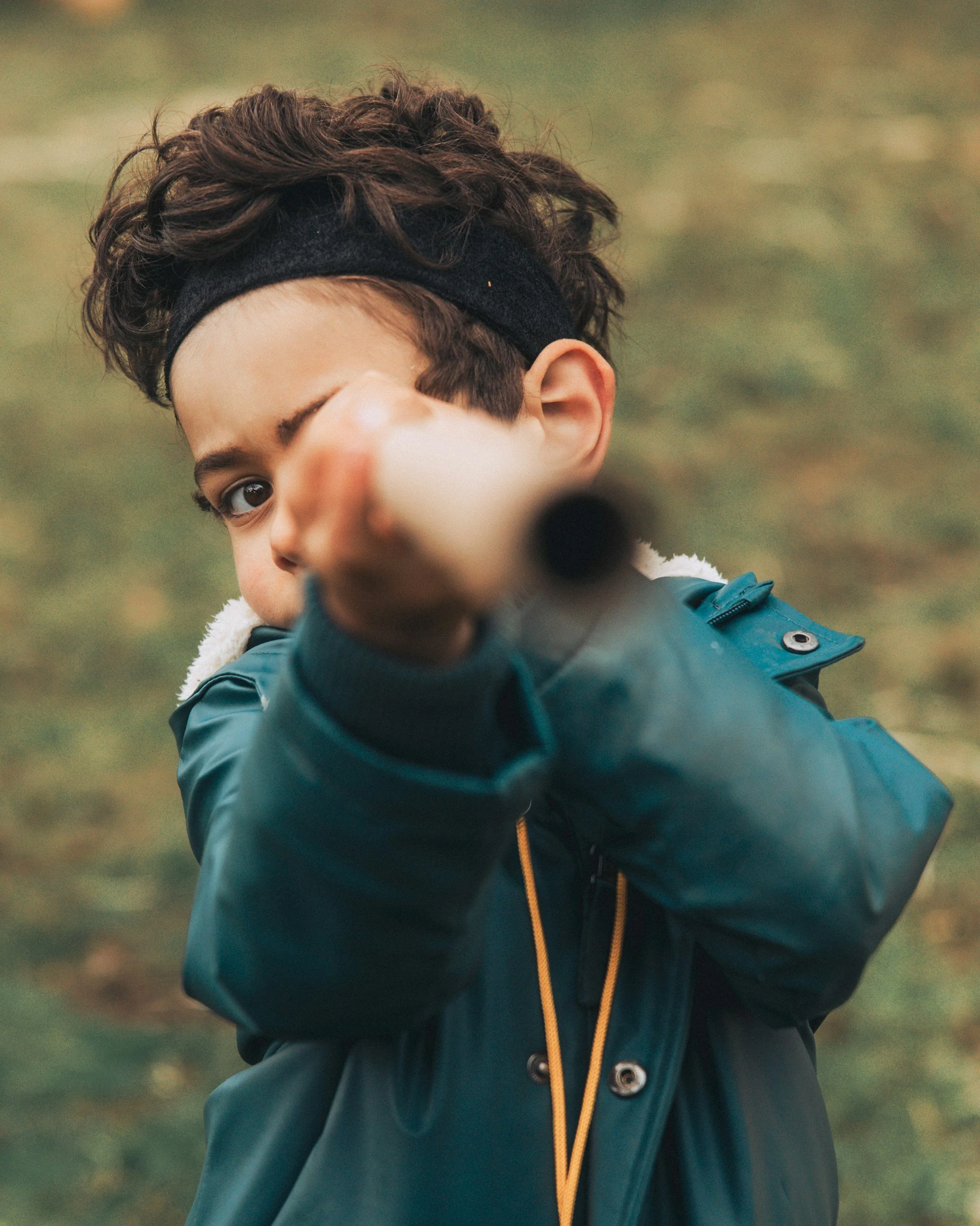 A young boy with curly brown hair, wearing a black headband and a teal jacket, points towards the camera with his right hand outdoors.