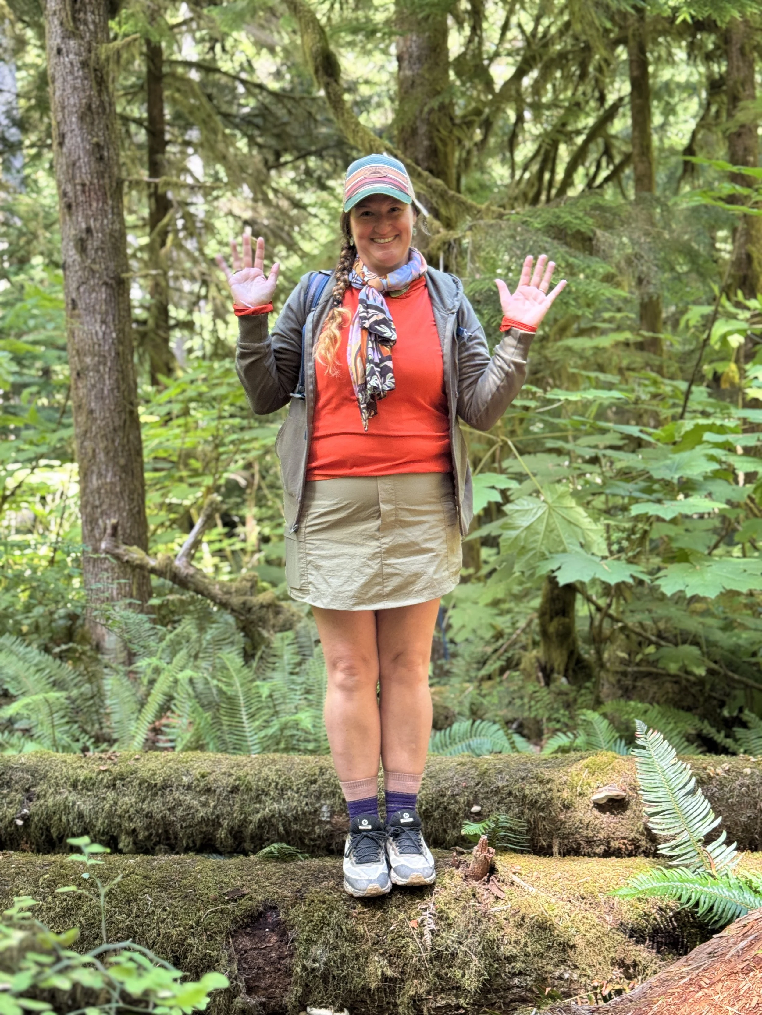 A woman stands on a moss-covered log in a lush green forest, smiling and waving at the camera. She is wearing a rainbow-striped baseball cap, a red shirt, a gray jacket, beige shorts, purple and gray socks, and sneakers. She has a backpack and a colorful scarf around her neck, with braided hair.