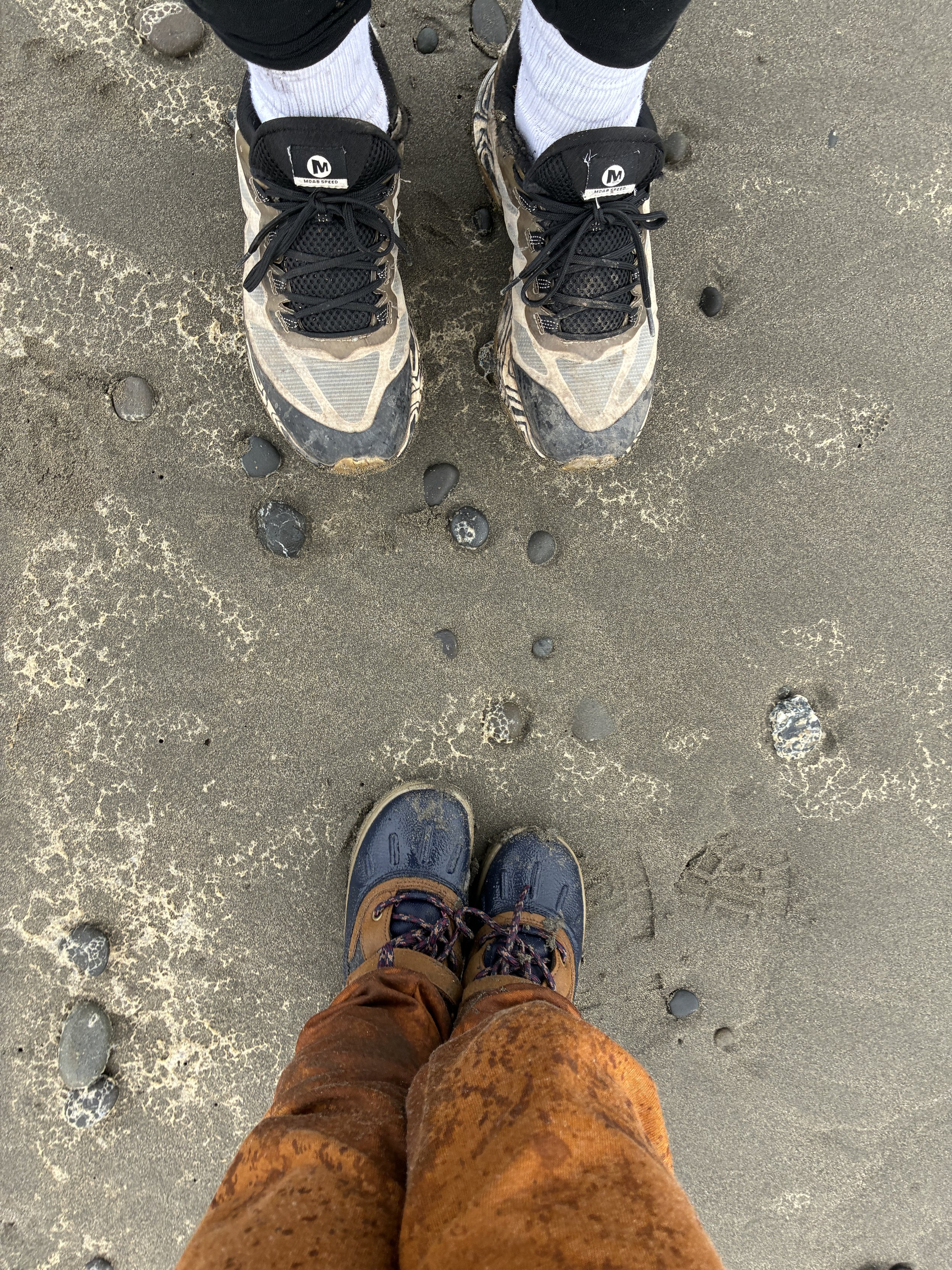 Two feet in hiking shoes standing on wet sand with scattered black stones and foam. The person is wearing orange-brown pants and brown hiking shoes with dark laces, and they are looking down at the surface.
