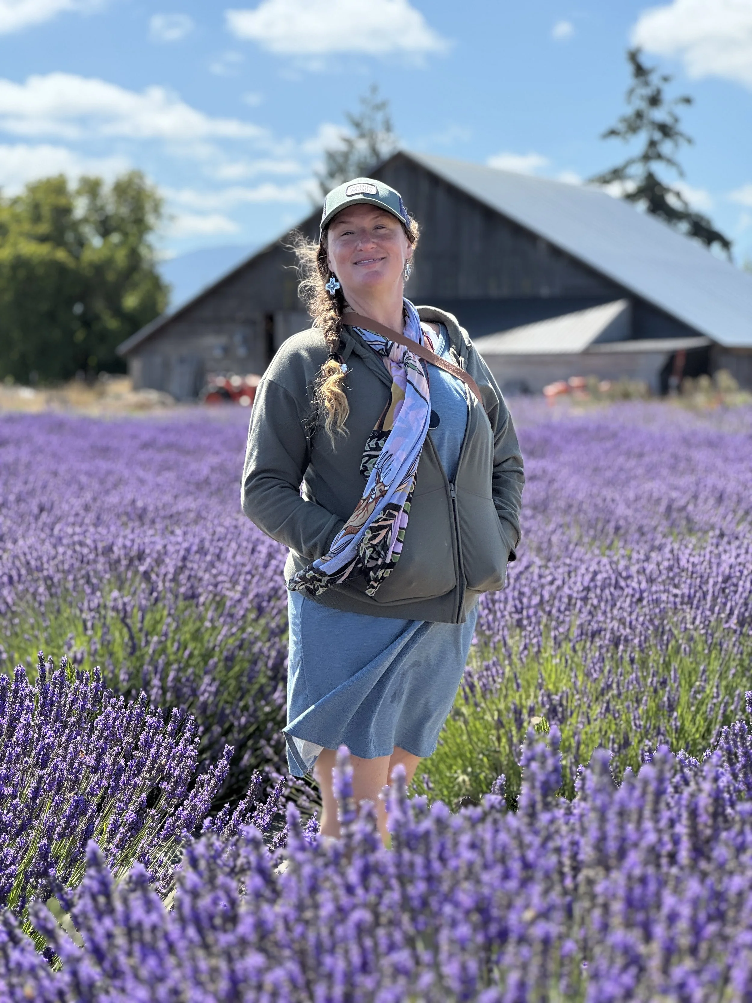 Woman standing in a field of purple lavender flowers, smiling, with a barn and trees in the background on a sunny day.