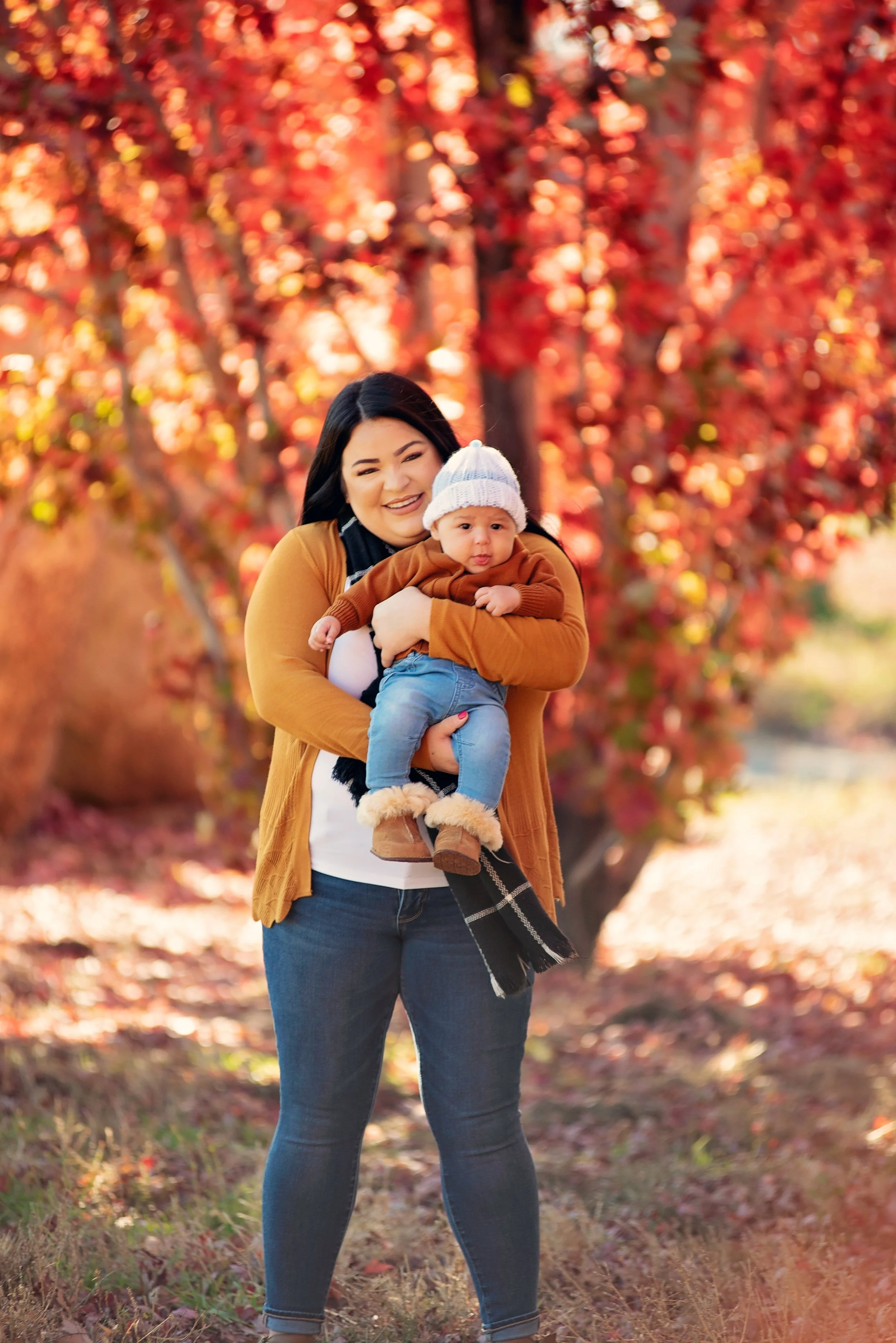 A woman with dark hair smiling and holding a baby in front of a background of autumn trees with red and orange leaves. It is daytime, and they are outdoors.