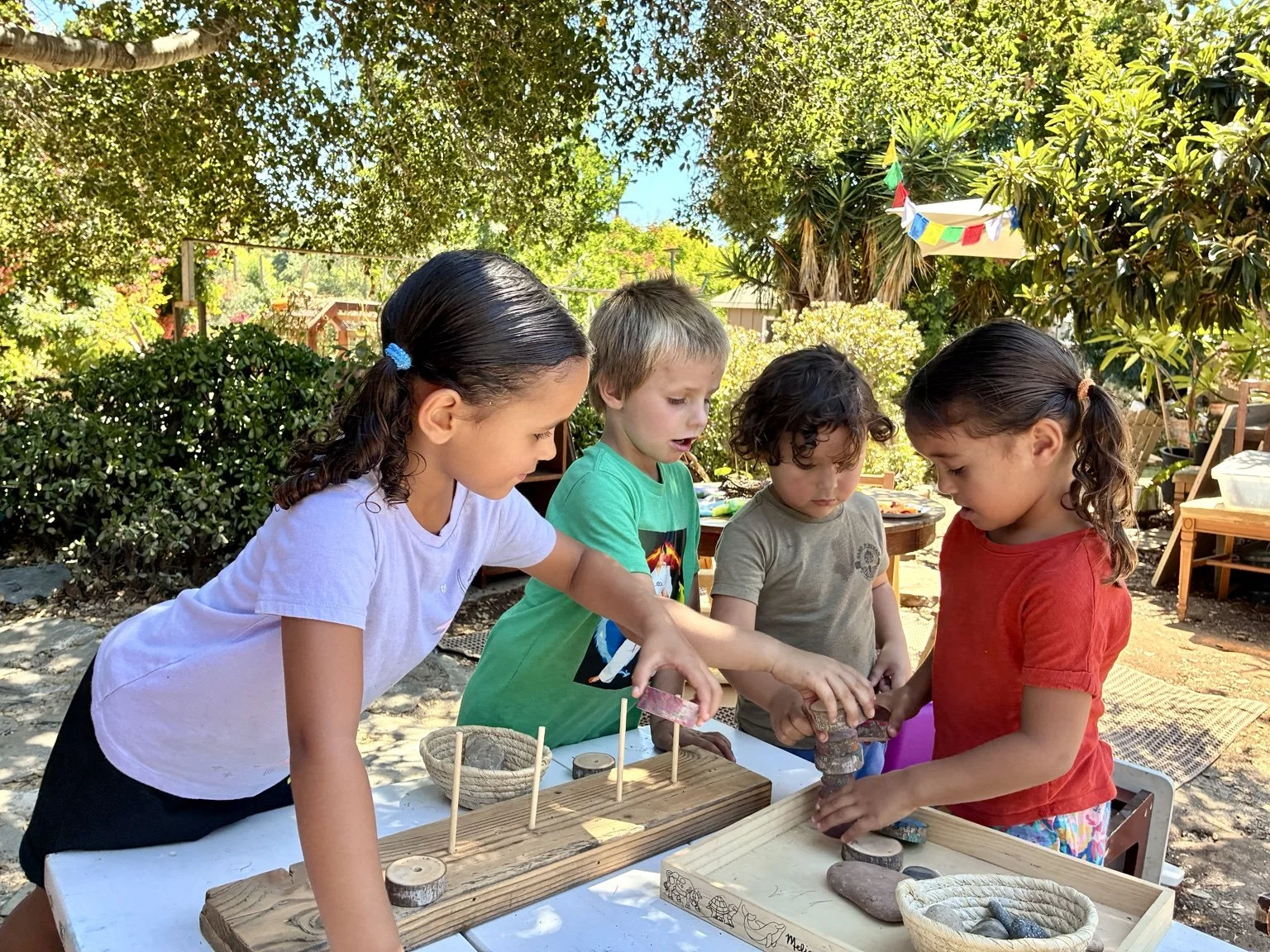Four children gather around an outdoor table, engaging with wooden and stone toys, amidst a lush garden setting with trees, bushes, and colorful decorations.