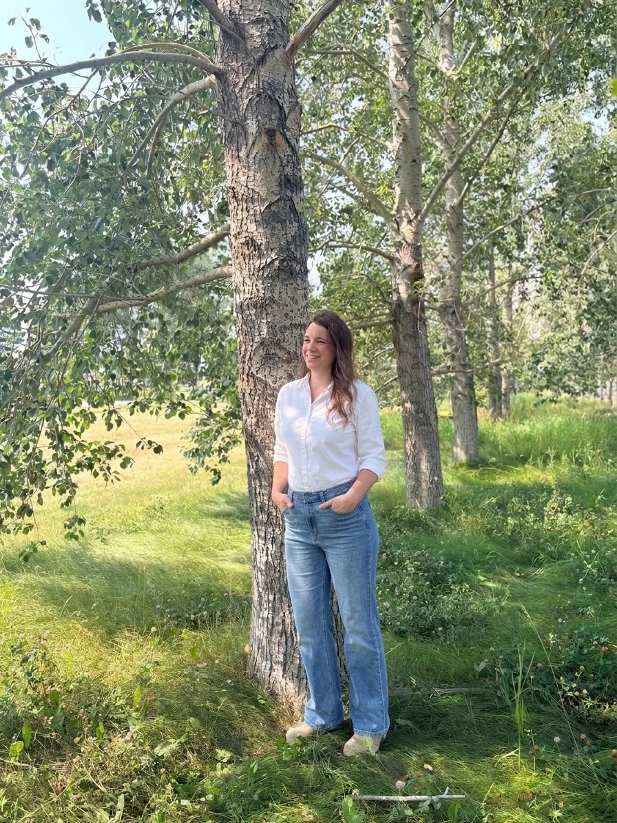 A woman standing outdoors near a large tree in a wooded area, smiling and looking to her left, wearing a white shirt and blue jeans.