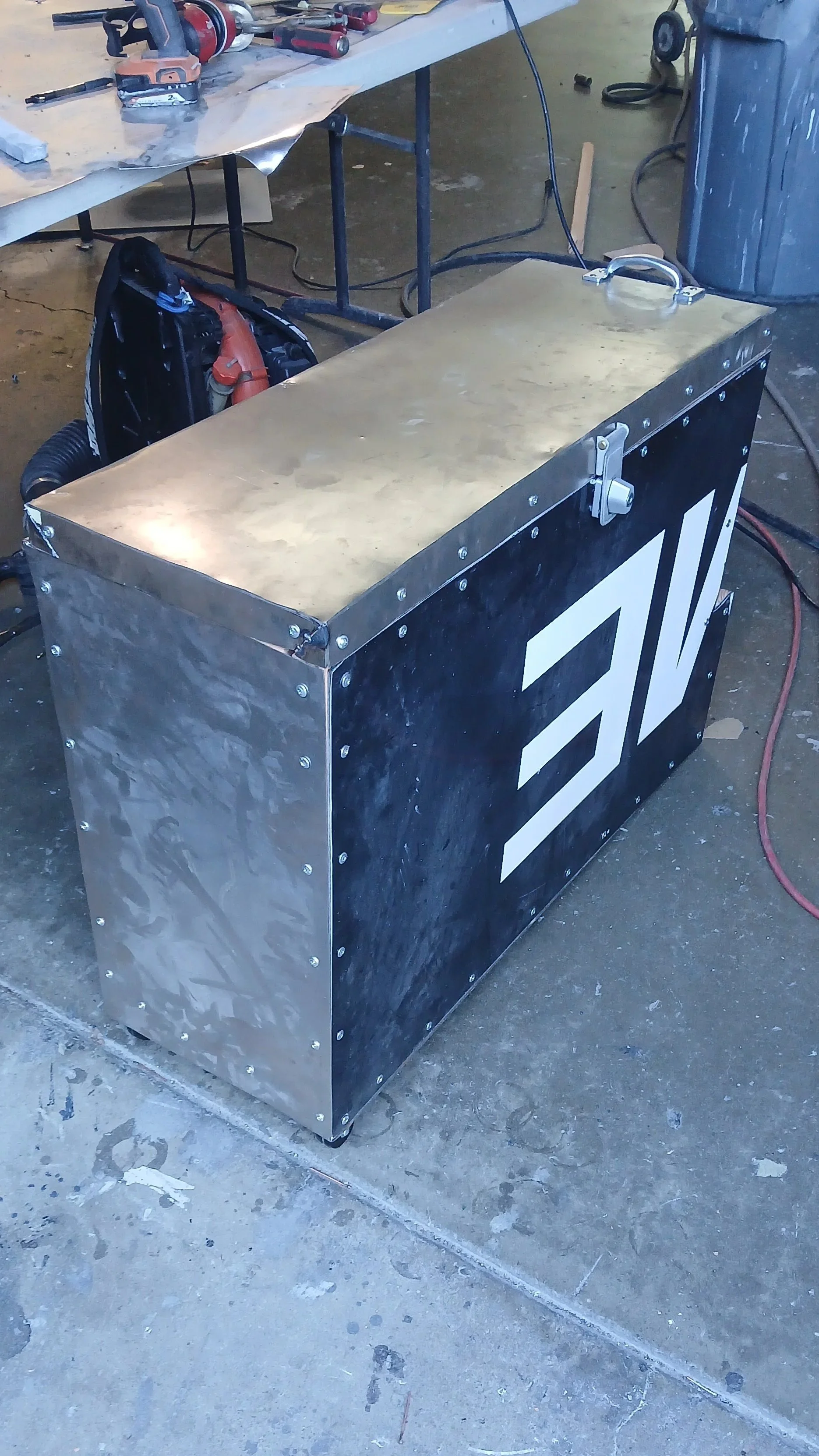 A metal welding cart with a black and white logo on the side, placed on a concrete floor in a workshop. The cart has a handle on top and an orange welding machine partially tucked underneath. In the background, there are workbenches, tools, and other equipment.