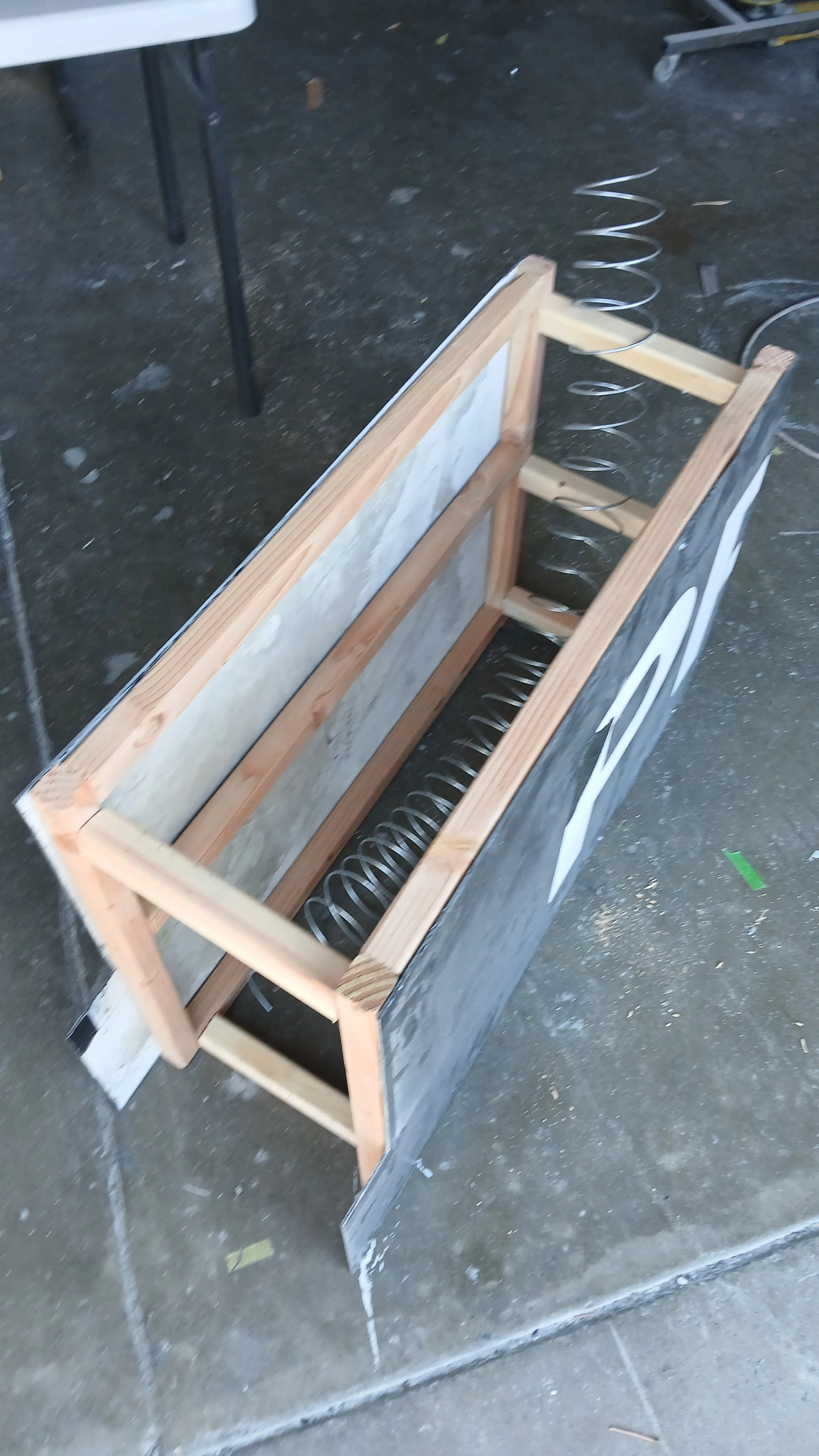 A wooden frame with a metal coil spring inside, likely a bed or chair frame under construction, on a concrete floor with scattered debris. First prototype of a Steady Vending Company Disc golf vending machine