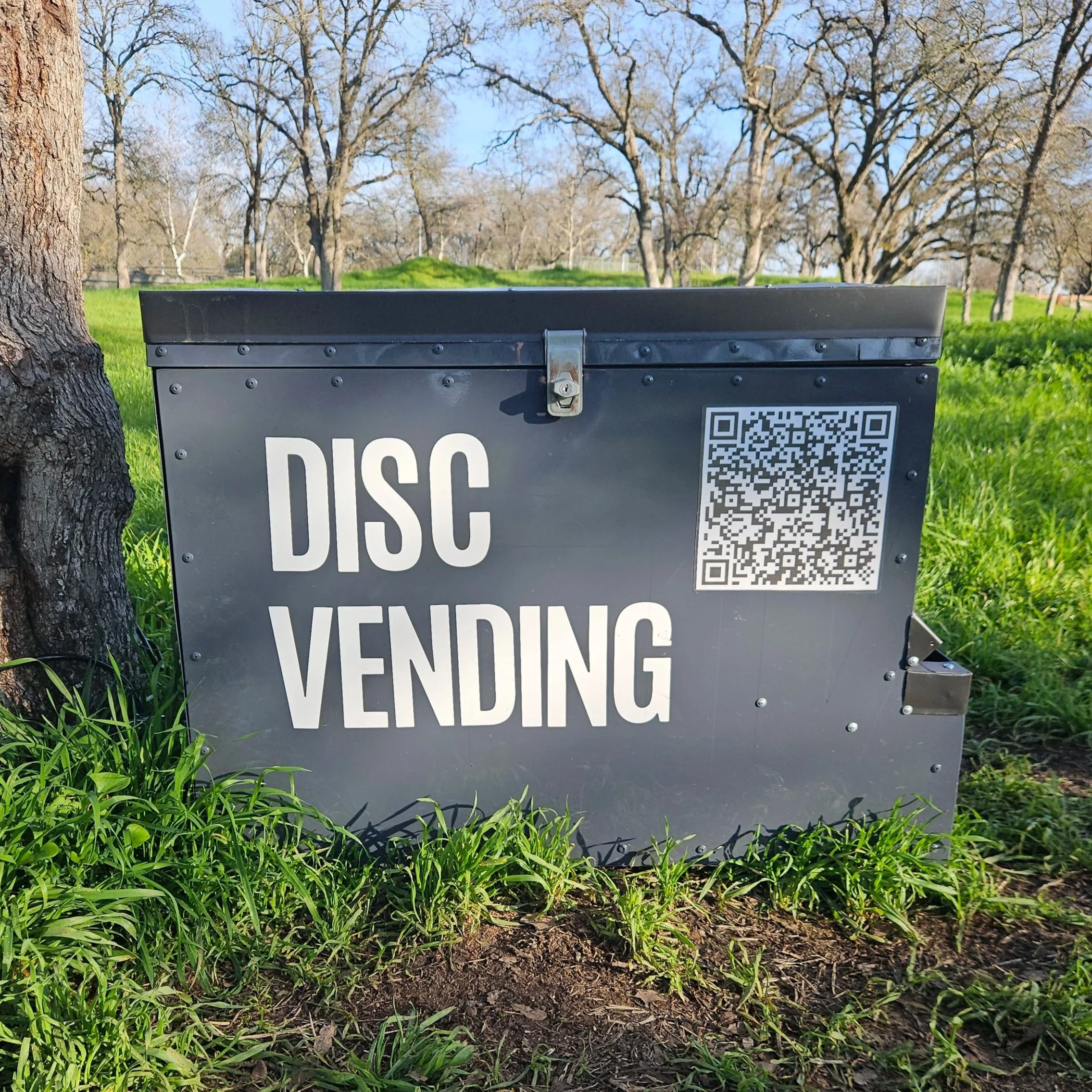 A disc golf vending machine with a QR code and partial text, situated in a park with green grass, trees, and blue sky in the background.