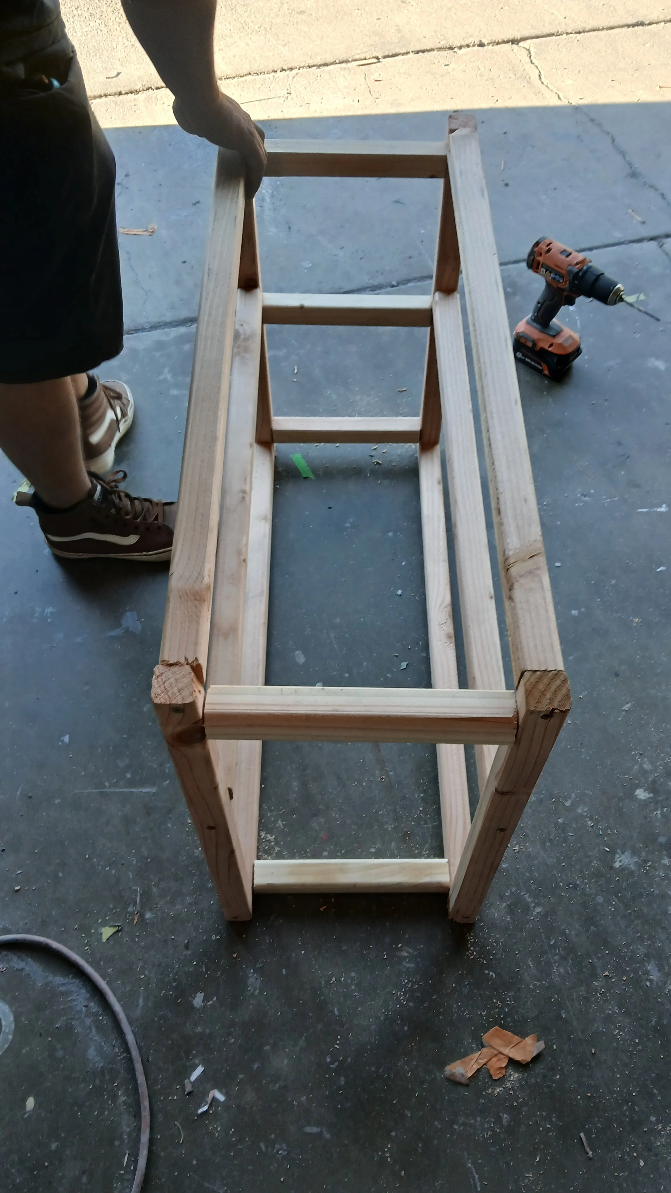 A wooden frame structure being built on a concrete floor next to a person wearing sneakers, with a cordless drill nearby and some small debris on the ground. Frame of a disc golf vending machine