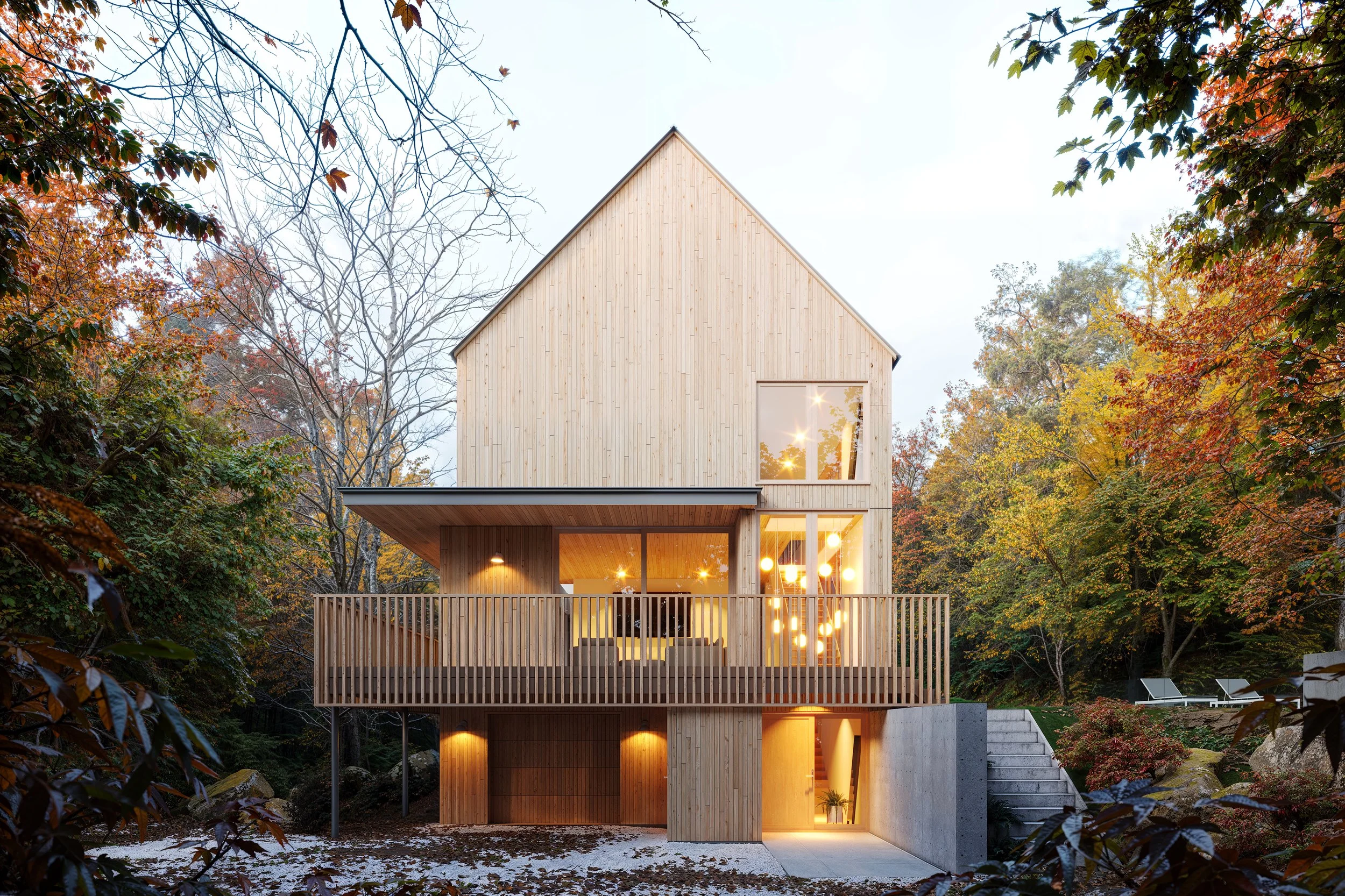 Contemporary timber house set within a lush autumn forest, featuring a minimalist gabled façade, warm natural wood cladding, large windows, a covered terrace, and inviting interior lighting that blends modern architecture with the surrounding landsca