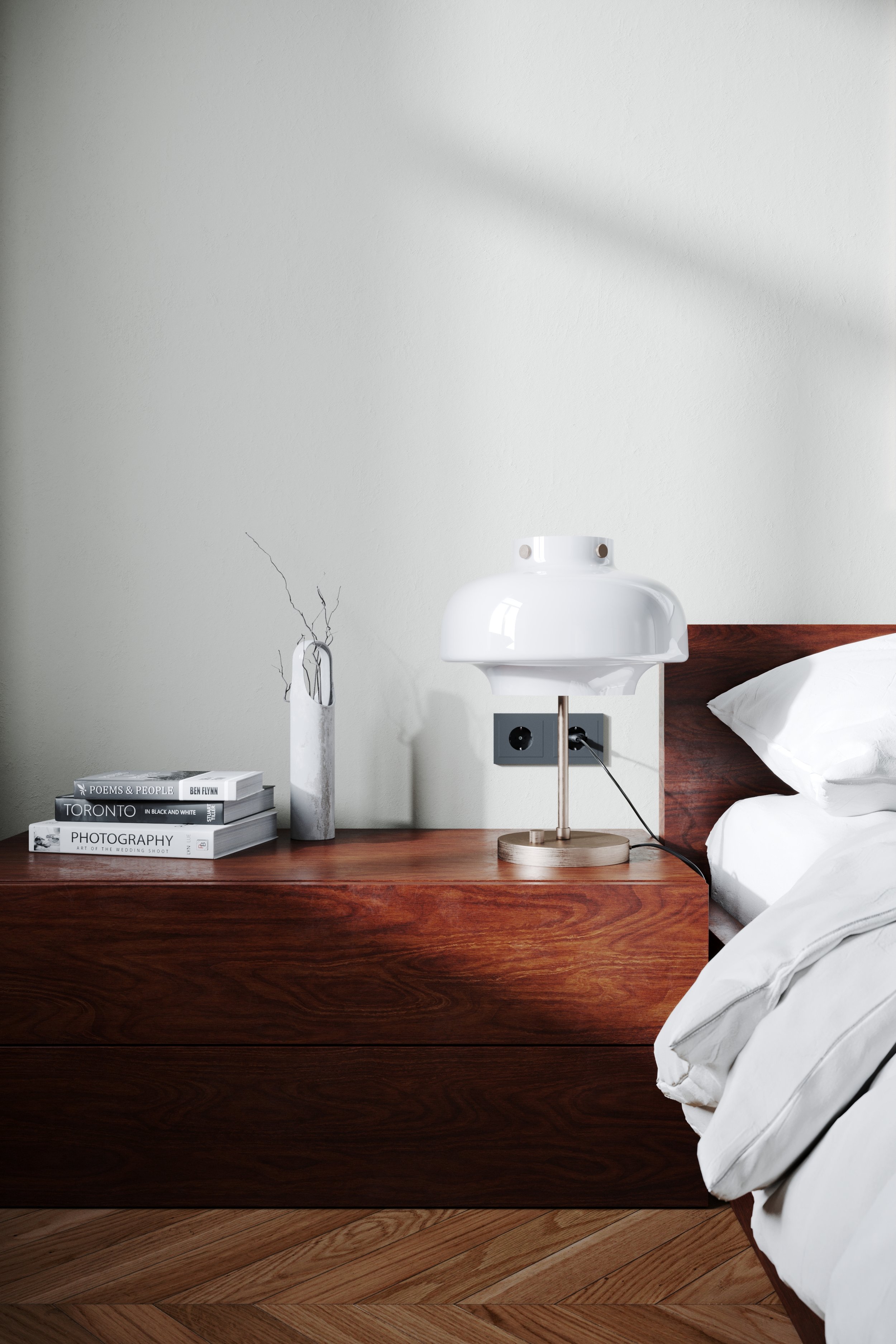 A wooden bed side table with a stack of books, a white vase with dried branches, and a white modern table lamp. Part of a bed with white linens is visible on the right. The wall behind is plain and light-colored. The floor is wooden.