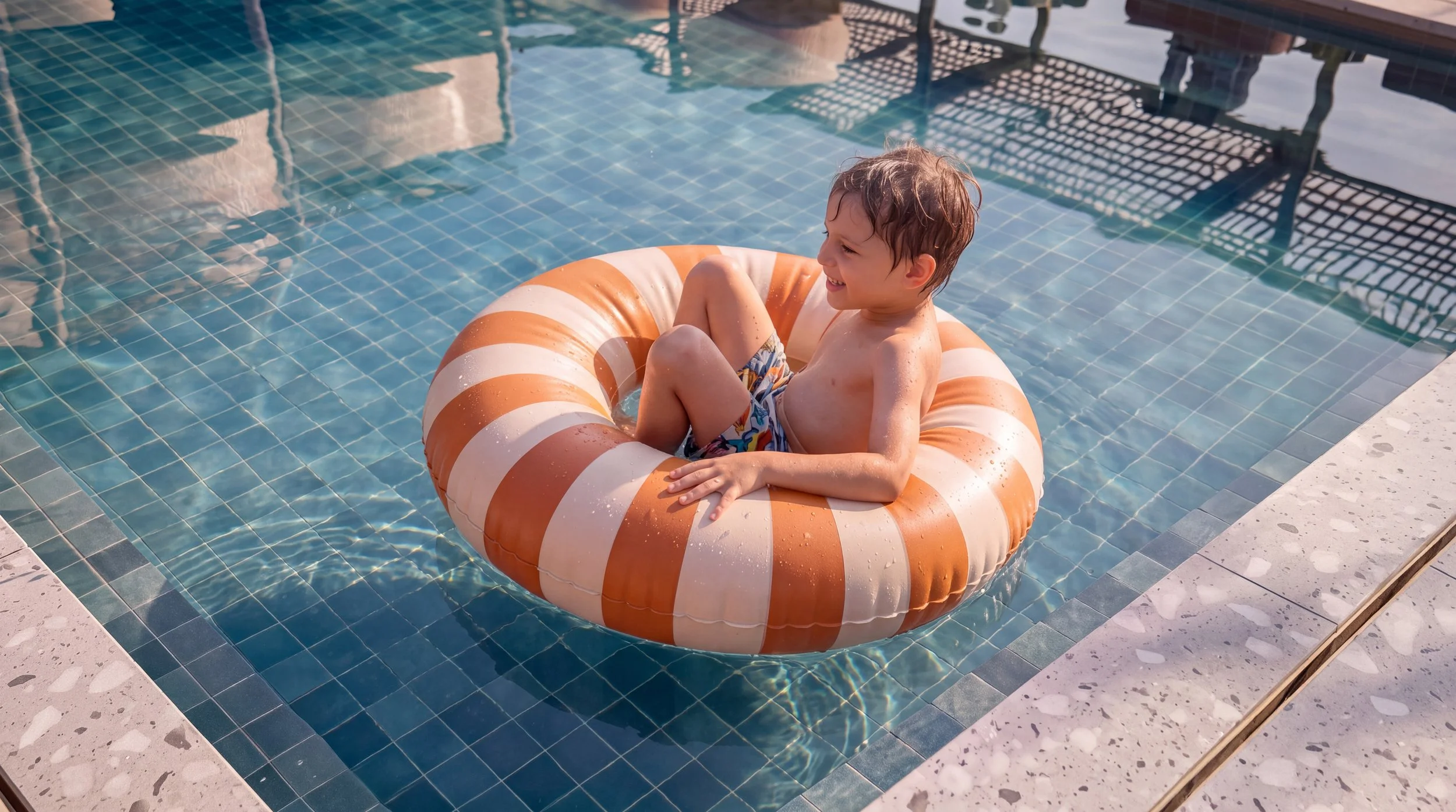 A young boy sitting in an orange and white striped inflatable pool float in a swimming pool, smiling and enjoying himself.
