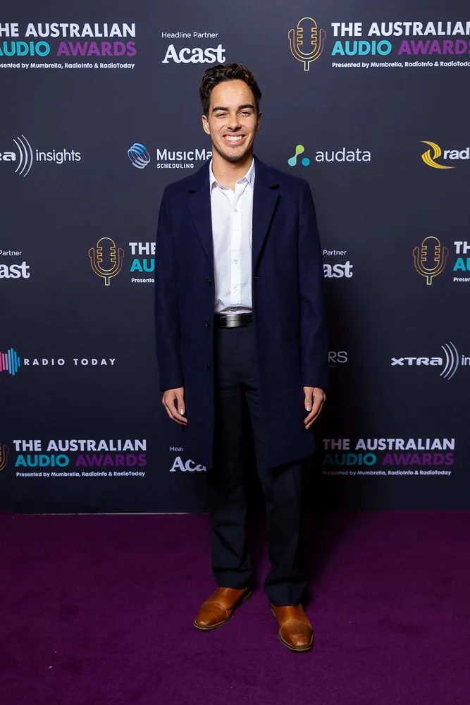 A young man in a dark blue overcoat, white shirt, black pants, and brown shoes standing on a purple carpet at the Australian Audio Awards event. He is smiling for the camera with a backdrop featuring sponsor logos and event branding.
