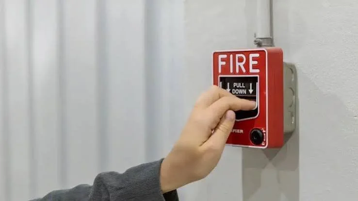 A person pulling down a fire alarm pull station on a wall.