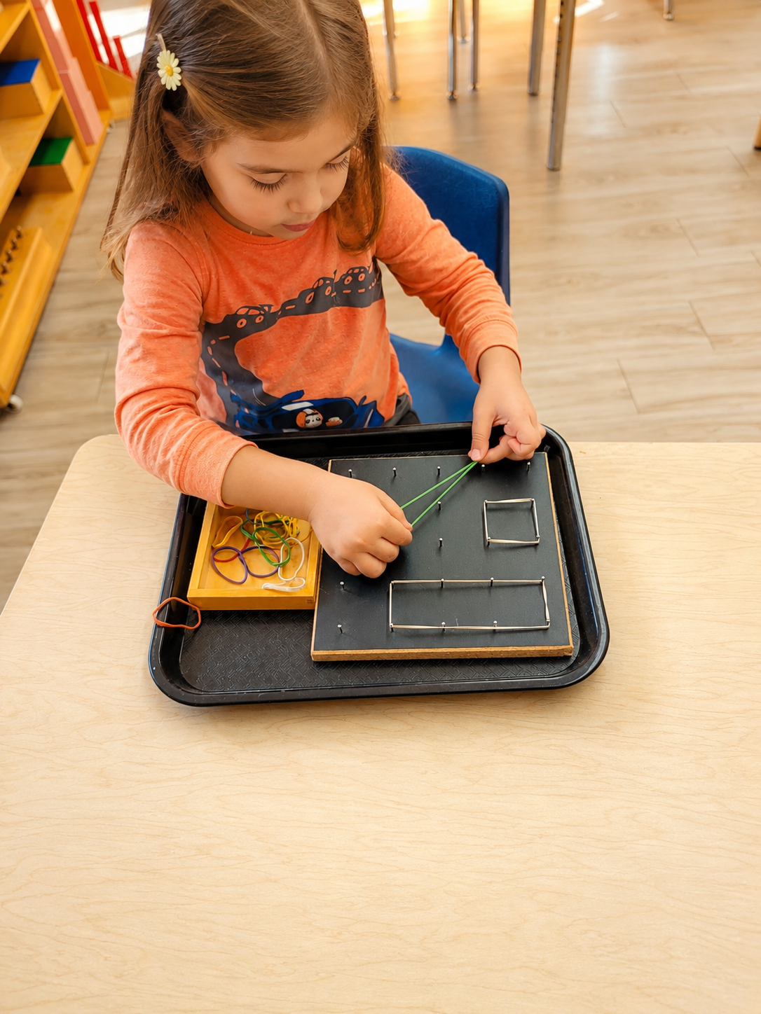 A young girl sitting at a table in a classroom, playing with geometric shape game pieces and rubber bands on a black board.