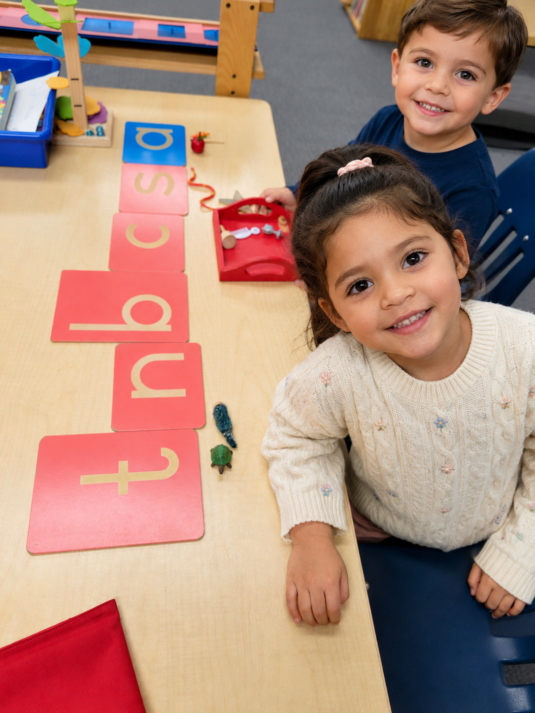 Two children, a girl and a boy, sitting at a classroom table with a row of letter cards spelling 't u p s a d.' The girl has curly hair, wearing a cream sweater, smiling at the camera. The boy has straight hair, wearing a navy blue shirt, smiling at the camera. The table has educational toys and materials.