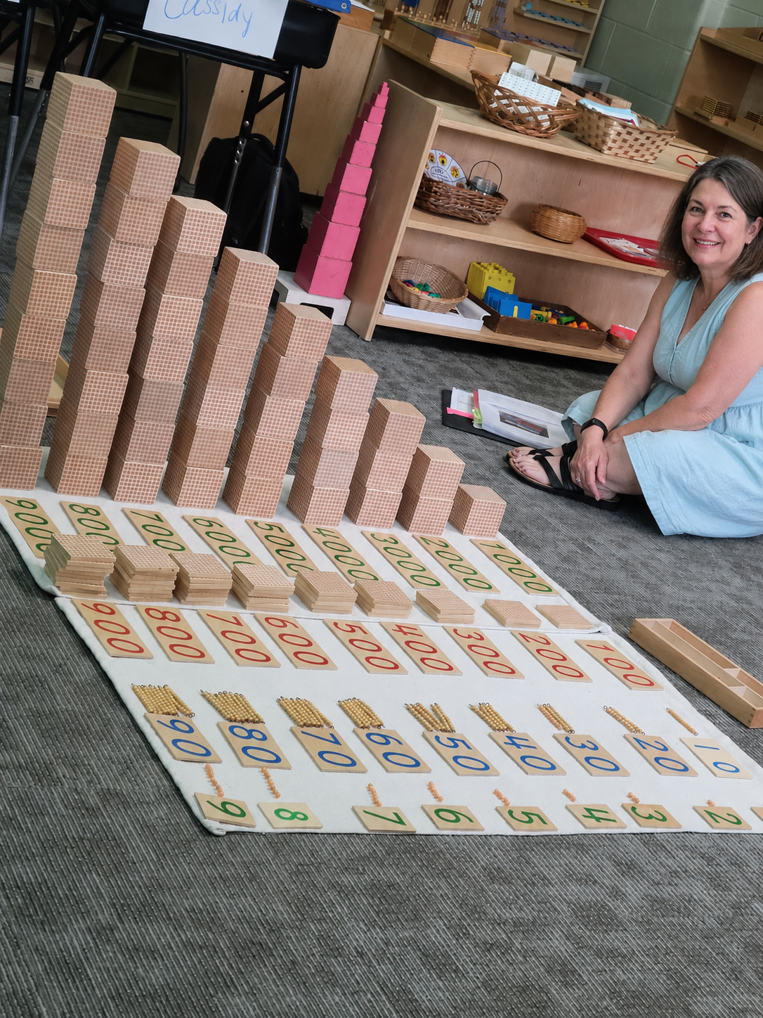 Educational display with model blocks, charts, and number cards arranged on the floor, with a woman seated nearby, in a classroom setting.