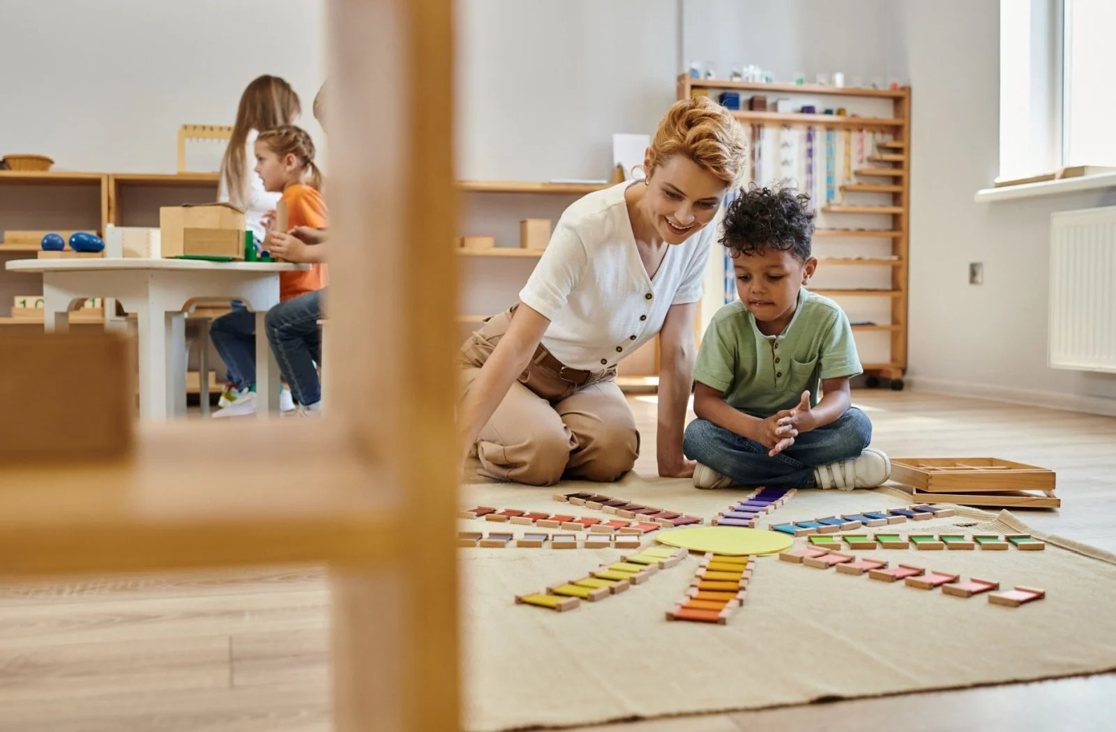 A teacher and young boy playing with colorful wooden tiles on the floor of a classroom, with other children in the background.