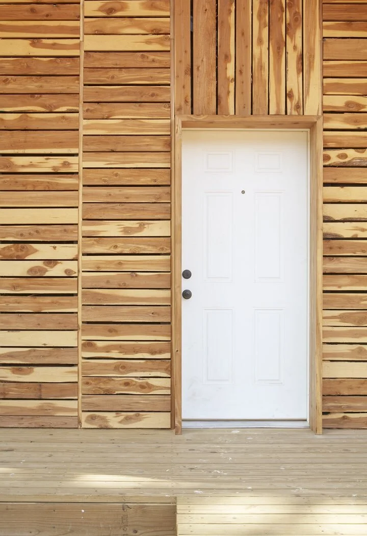 A white door with a peephole, set in a wooden wall made of horizontal planks and vertical slats.