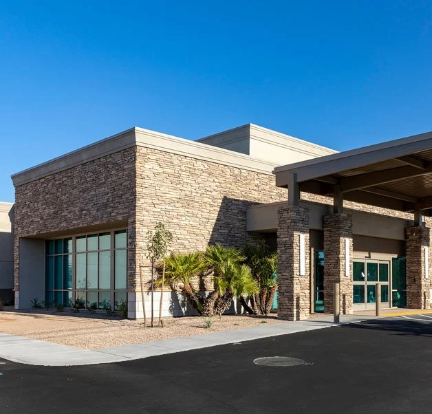 Exterior of a modern commercial building with stone and stucco walls, large windows, palm trees, and a covered entrance, under a clear blue sky.