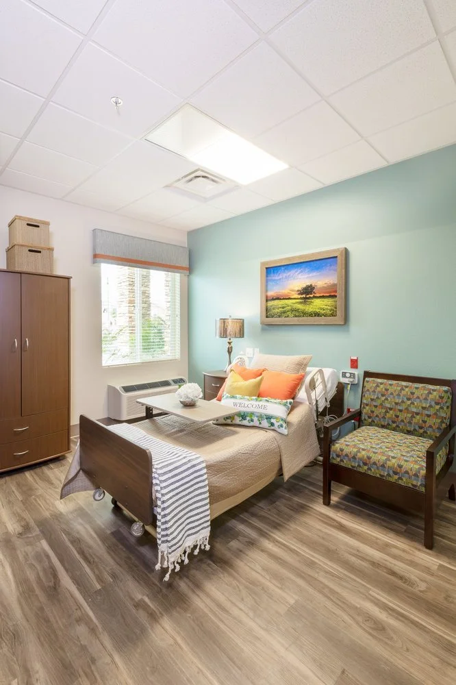 A hospital bedroom with a bed, a chair, a nightstand with a lamp, a painting on the wall, a window with blinds, and wooden flooring.