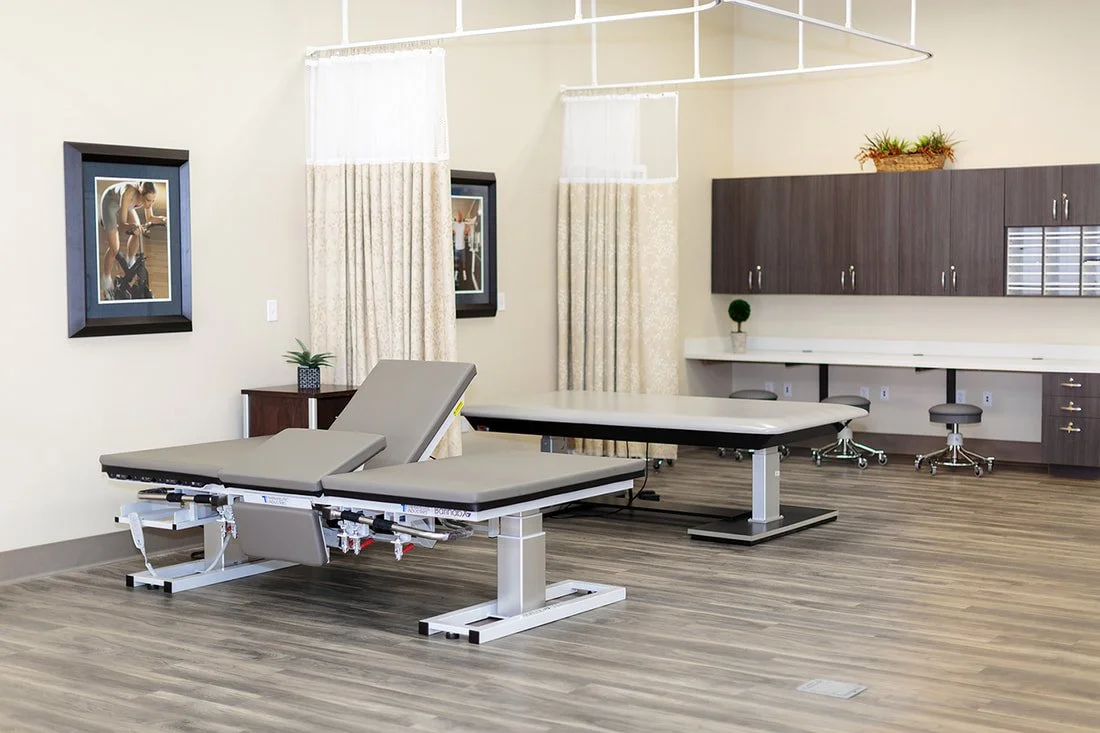 Empty medical examination room with an adjustable examination table, curtains, framed pictures, and medical cabinets.