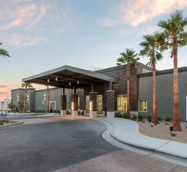 Modern commercial building with a covered entrance, palm trees, and a spacious parking area at sunset.