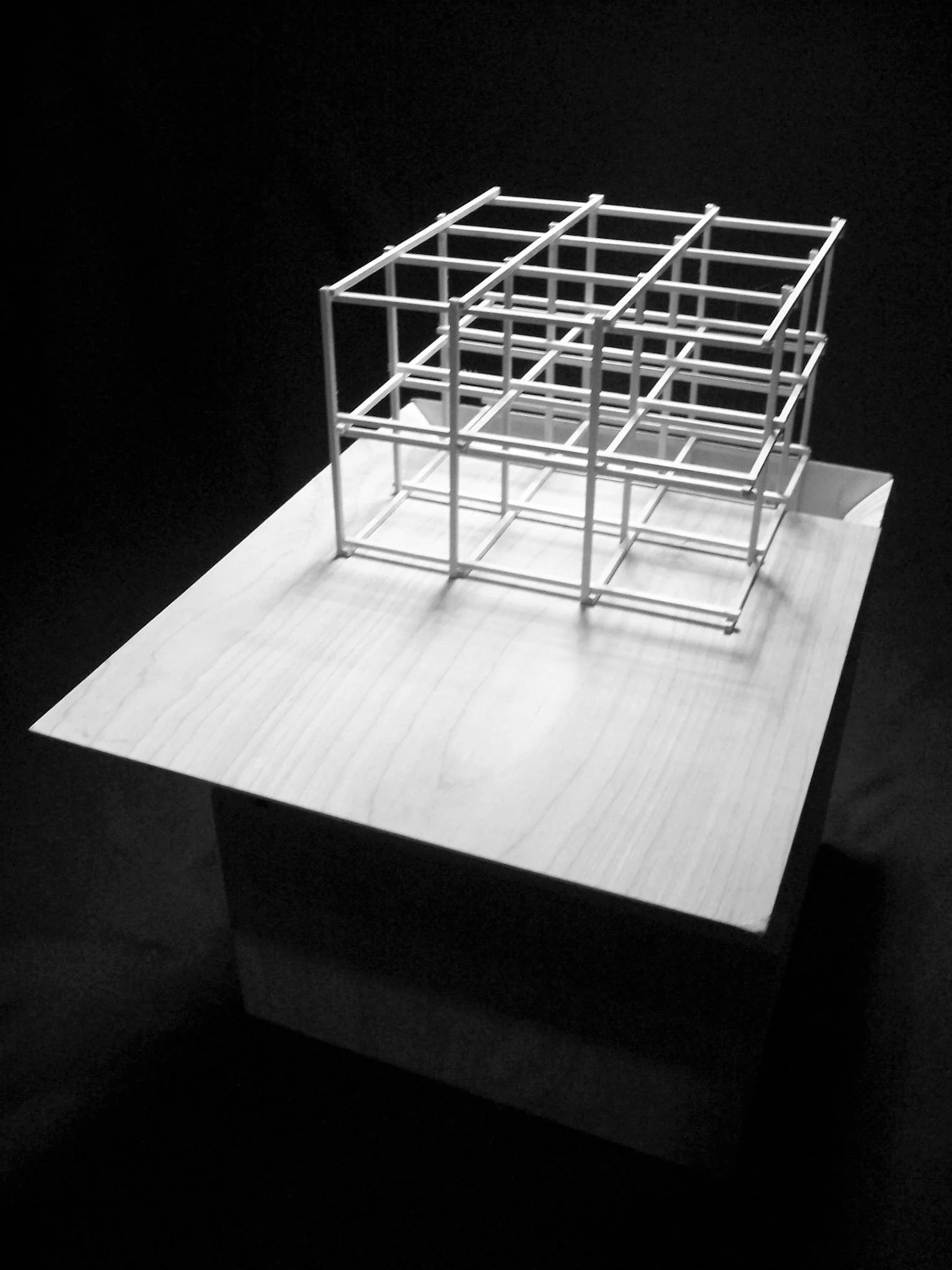 Minimalist architectural model of a multi-level building with a grid-like structure, placed on a wooden base against a black background.