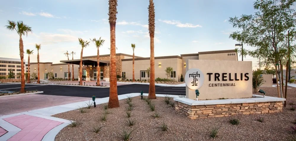 Exterior view of a modern building housing Trellis Centennial, with palm trees, landscaping, and a sign displaying the name and logo.