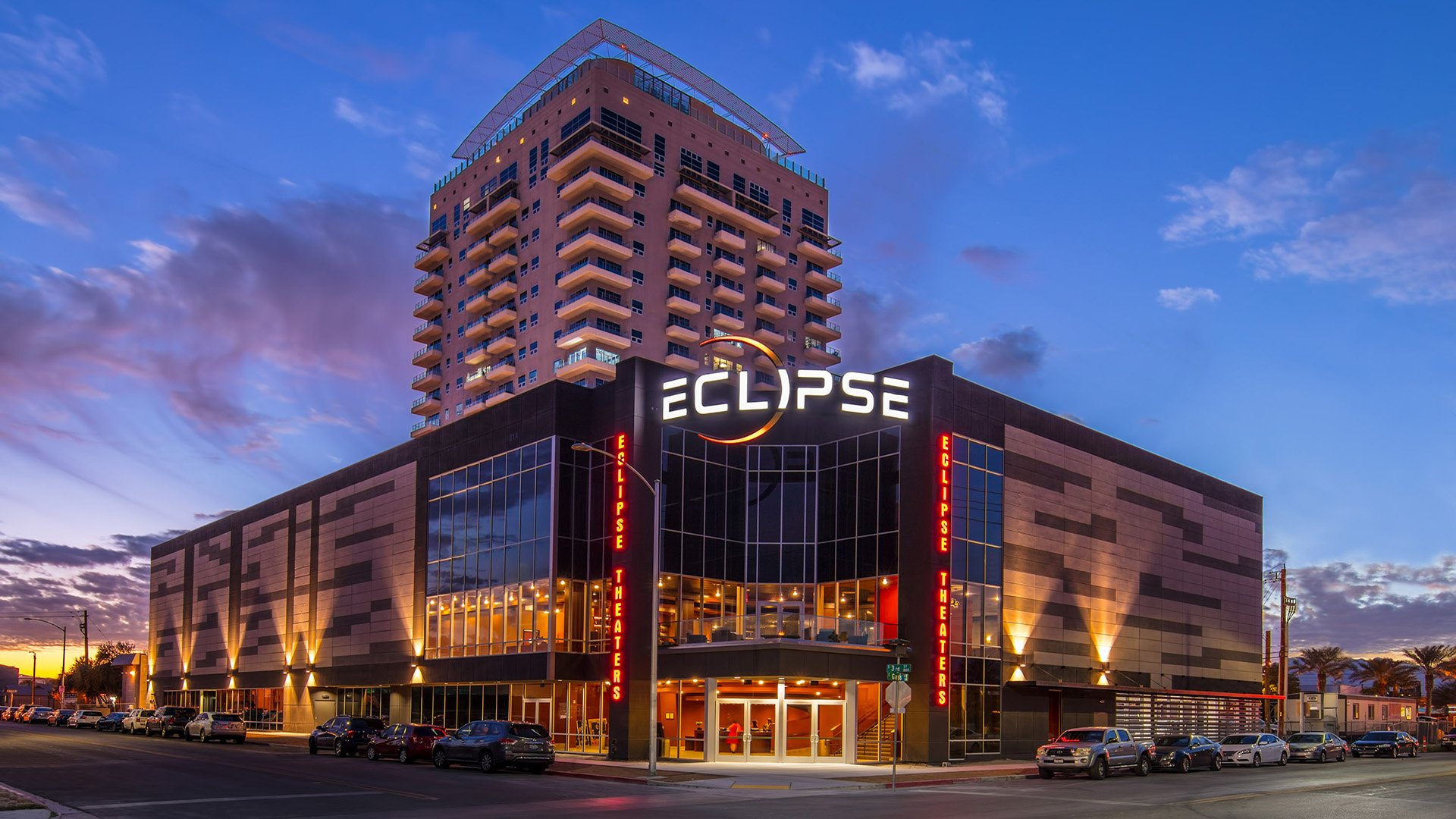 Night view of Eclipse Theaters with illuminated signs, a multi-story building with a tall stage tower, glass windows, parked cars in front, and a partly cloudy sky in the background.