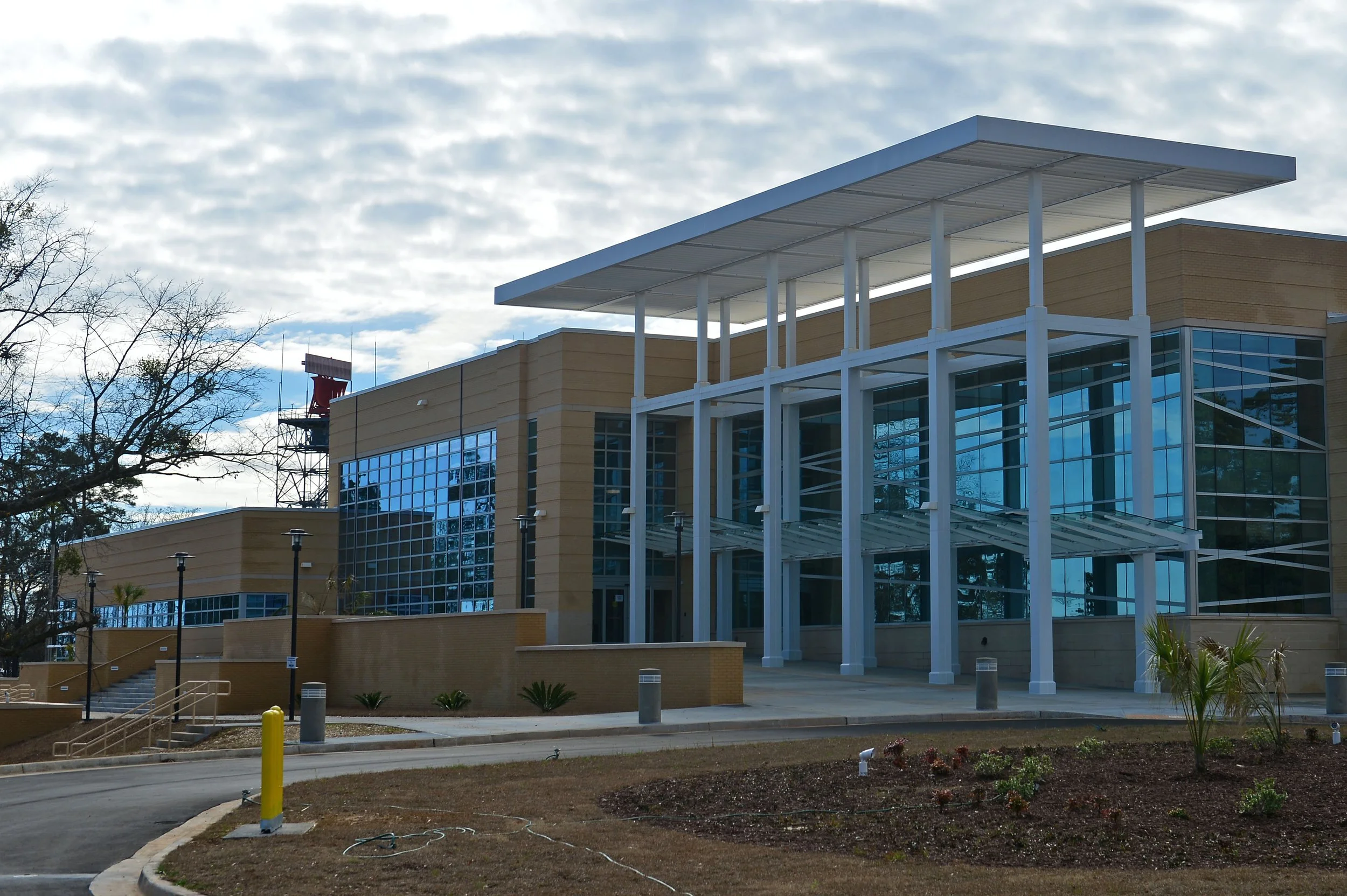 Modern building with large glass windows and a white metal roof structure, surrounded by a paved area, landscaped with small plants, and a cloudy sky overhead.