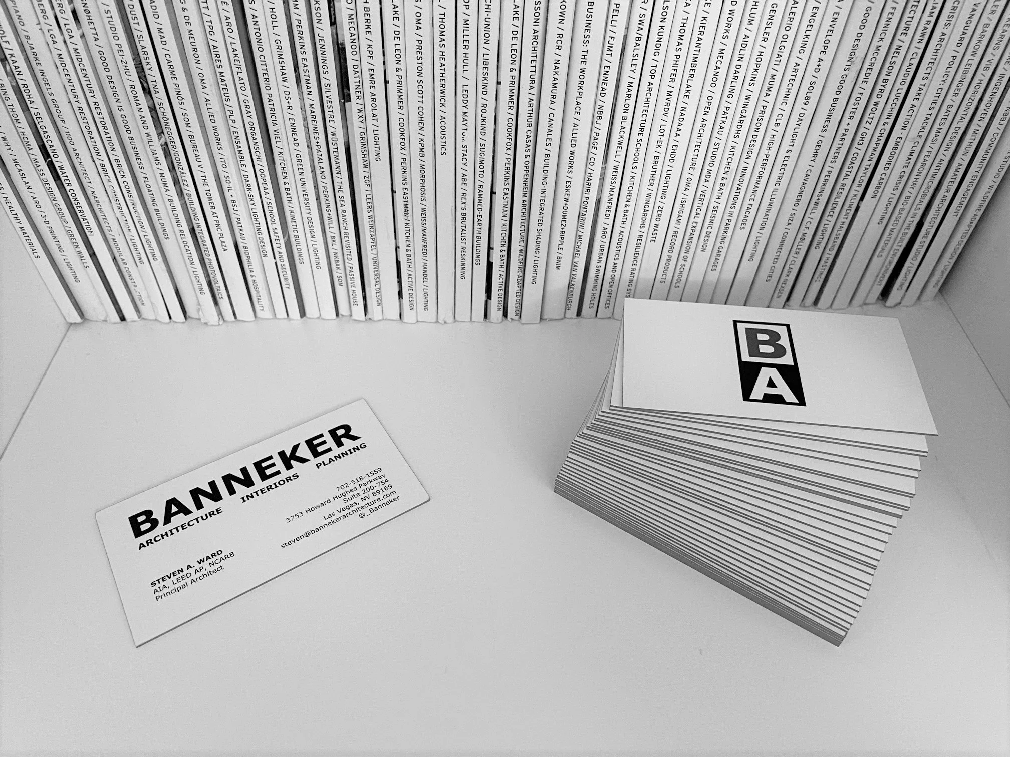 Close-up of a row of white business cards featuring bold black text that says 'BANNER ARCHITECTURE'. The cards rest on a white surface, with some cards stacked and others placed flat, and there are printed materials arranged around them.