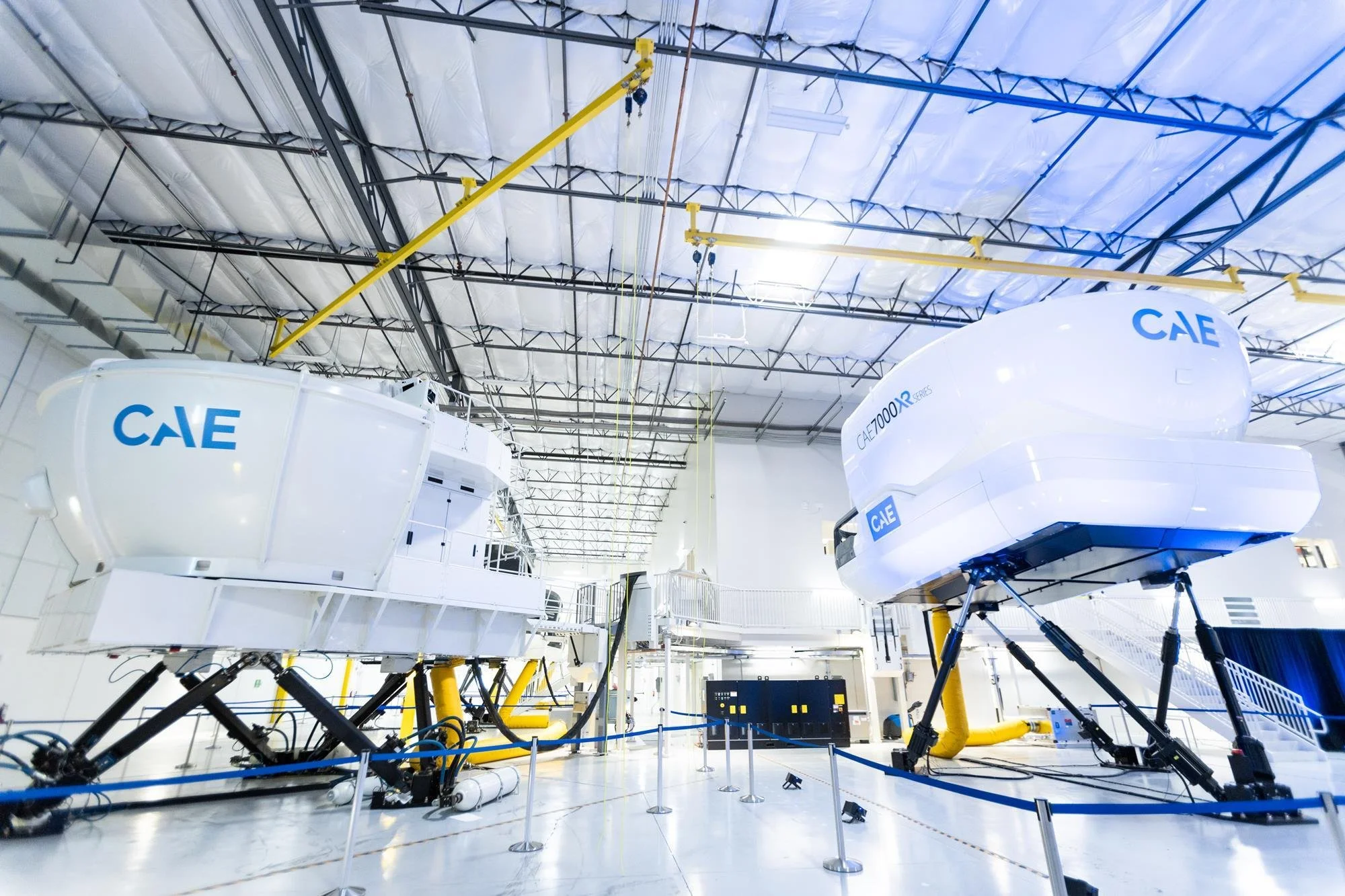 Inside a large facility with two white spacecraft labeled 'CAE' and 'CAE700XR SERIES', suspended by yellow hoists, with a high ceiling and industrial lighting.