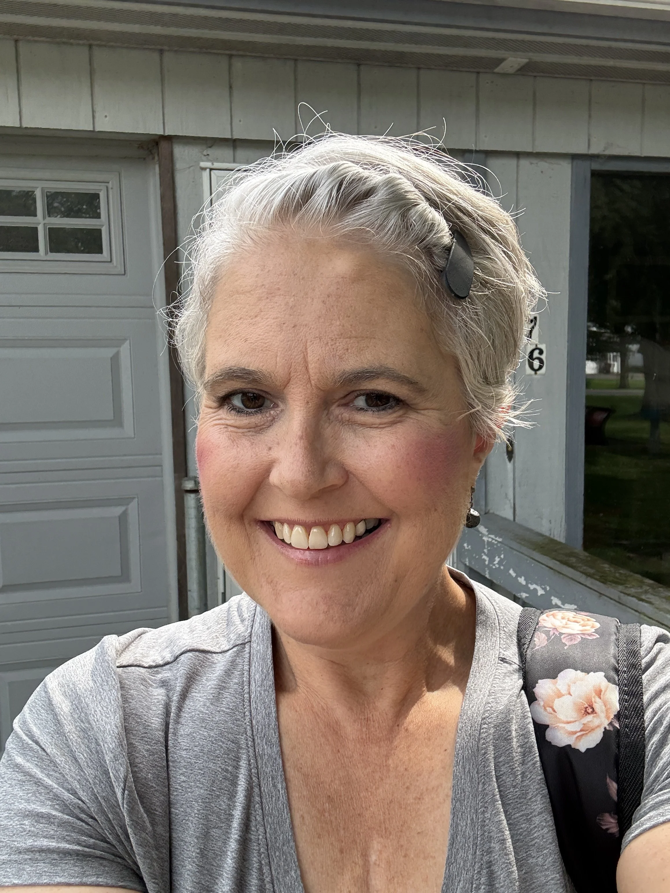A smiling woman with short gray hair, wearing a light gray shirt and a floral strap, standing outside near a garage and house with gray siding.