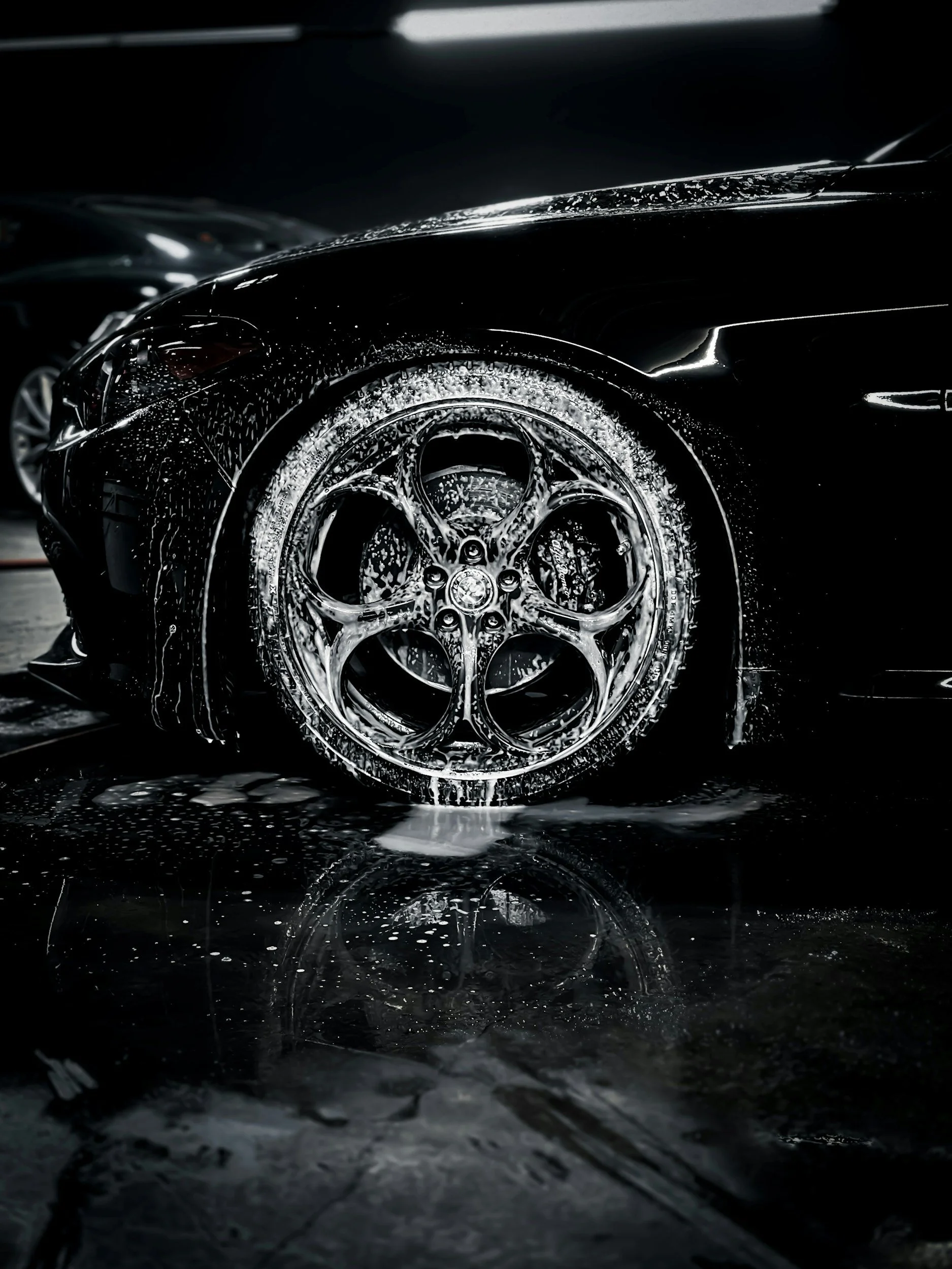 Close-up photo of a black sports car being washed, with water and soap suds on the tire and wheel, reflective surface and a dark background.
