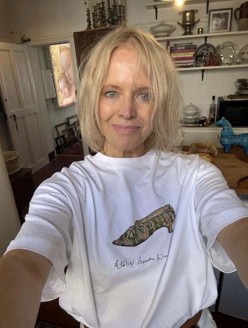A woman in a cozy, well-decorated room. She has blonde, shoulder-length, wavy hair and is wearing a white t-shirt with an original artwork, Provence design of a slipper and the words "Atelier Amanda Daniel" underneath.