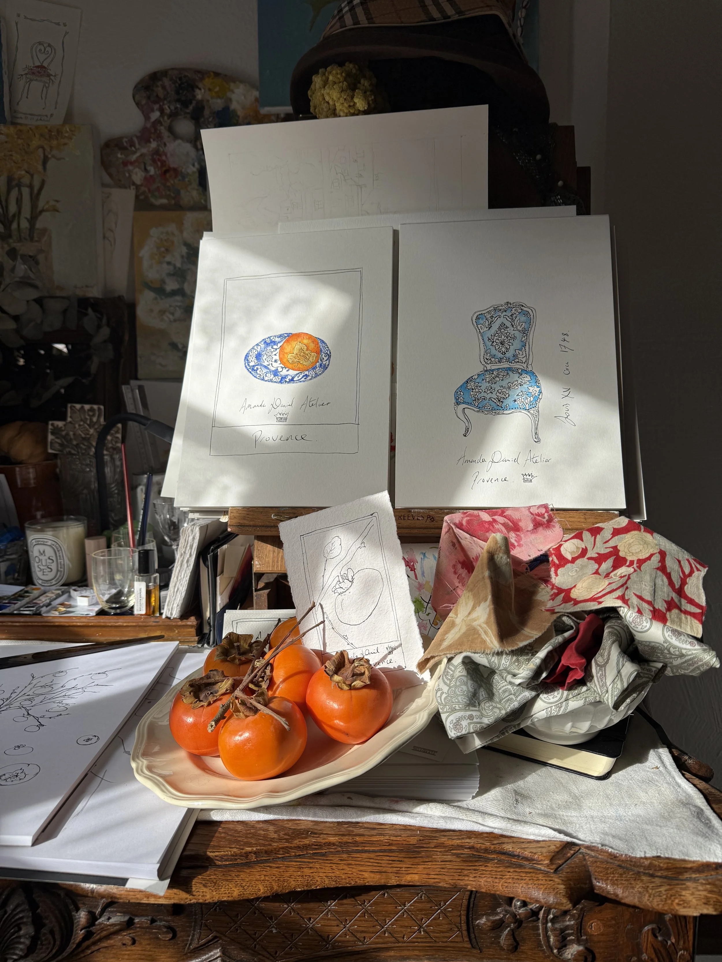 A wooden artists table with a white tablecloth displaying clear glass vases and orange persimmons on a decorative ceramic tray. The background has sketches, art supplies, and fabric, suggesting an art studio or workspace.