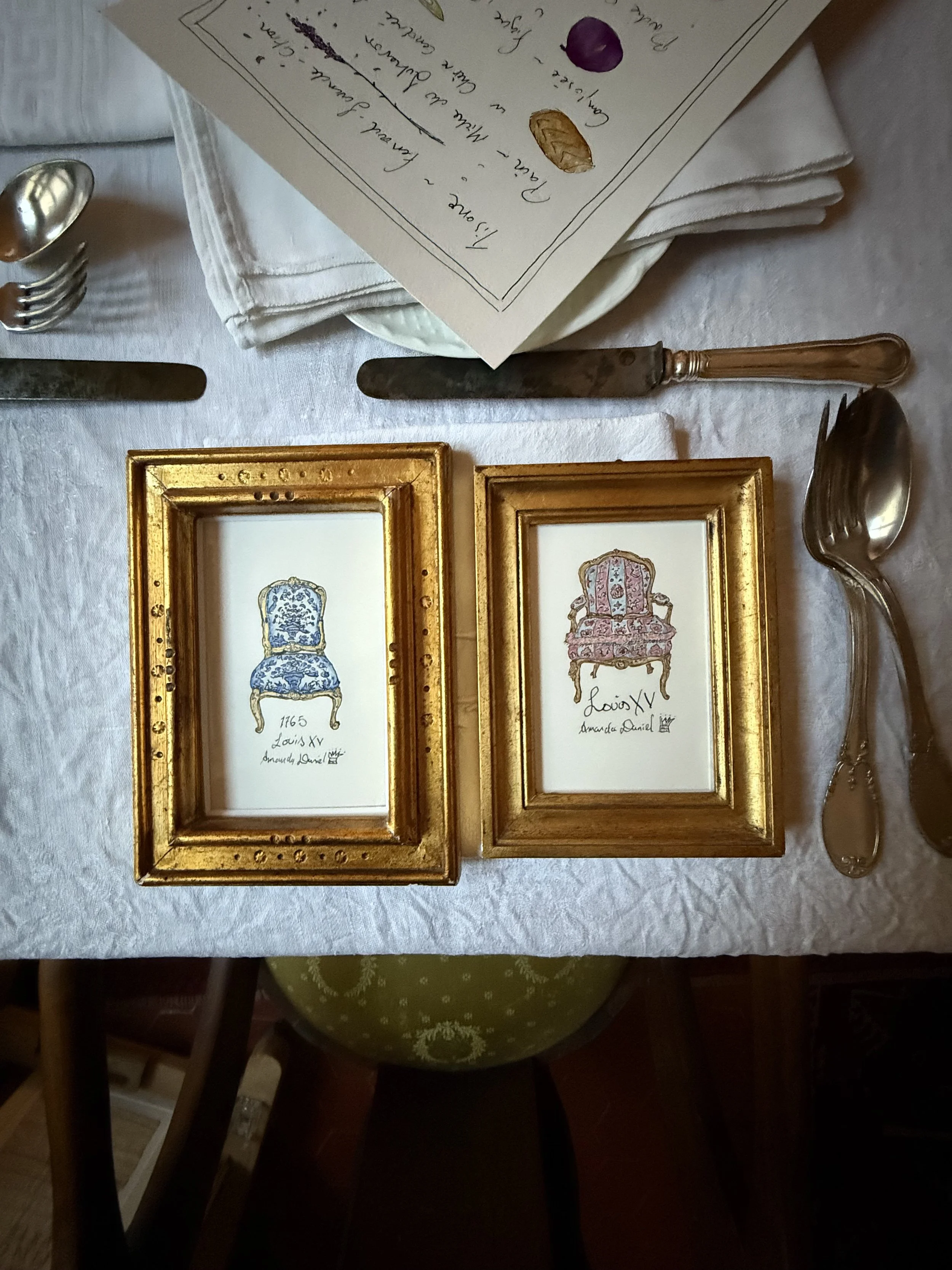 A place setting at a table featuring folded white cloth napkins, a menu, and framed pictures of ornate chairs with gold frames.