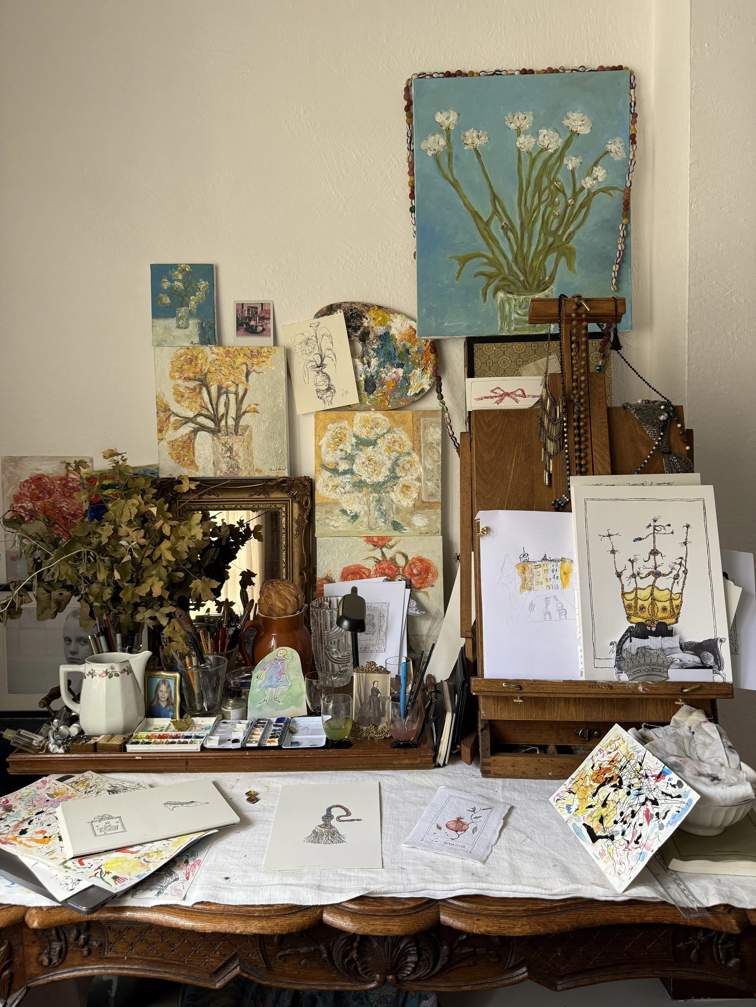 An artist's workspace with paintings of flowers on the wall, art supplies on the table, and sketches and paintings in progress.
