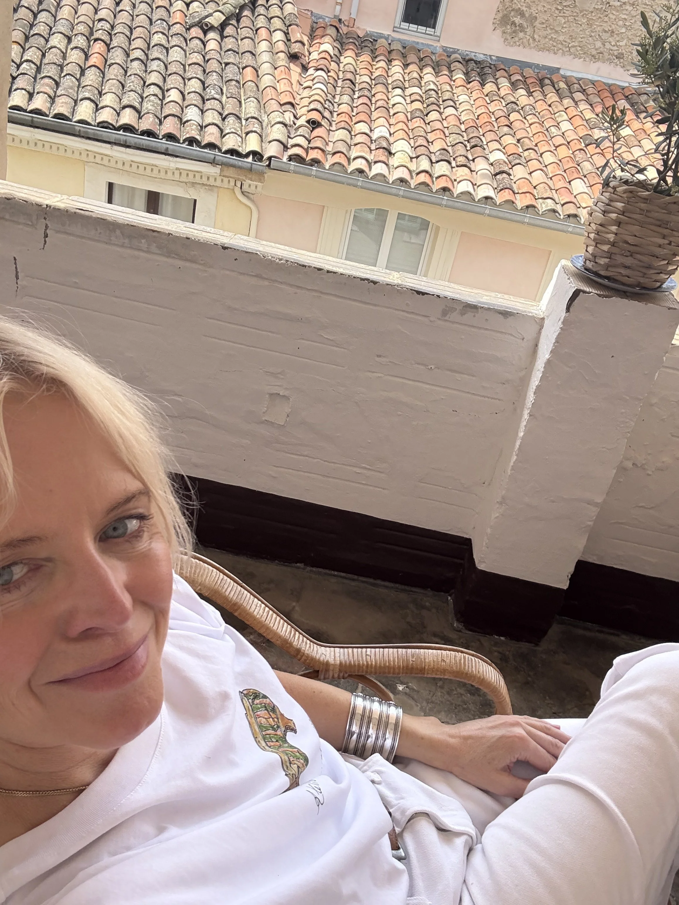 Close-up of a smiling woman with blonde hair, wearing a white shirt with a colorful Provence design, sitting outdoors in sunny weather. In the background, there's a terracotta-tiled roof and part of a building wall with a window.