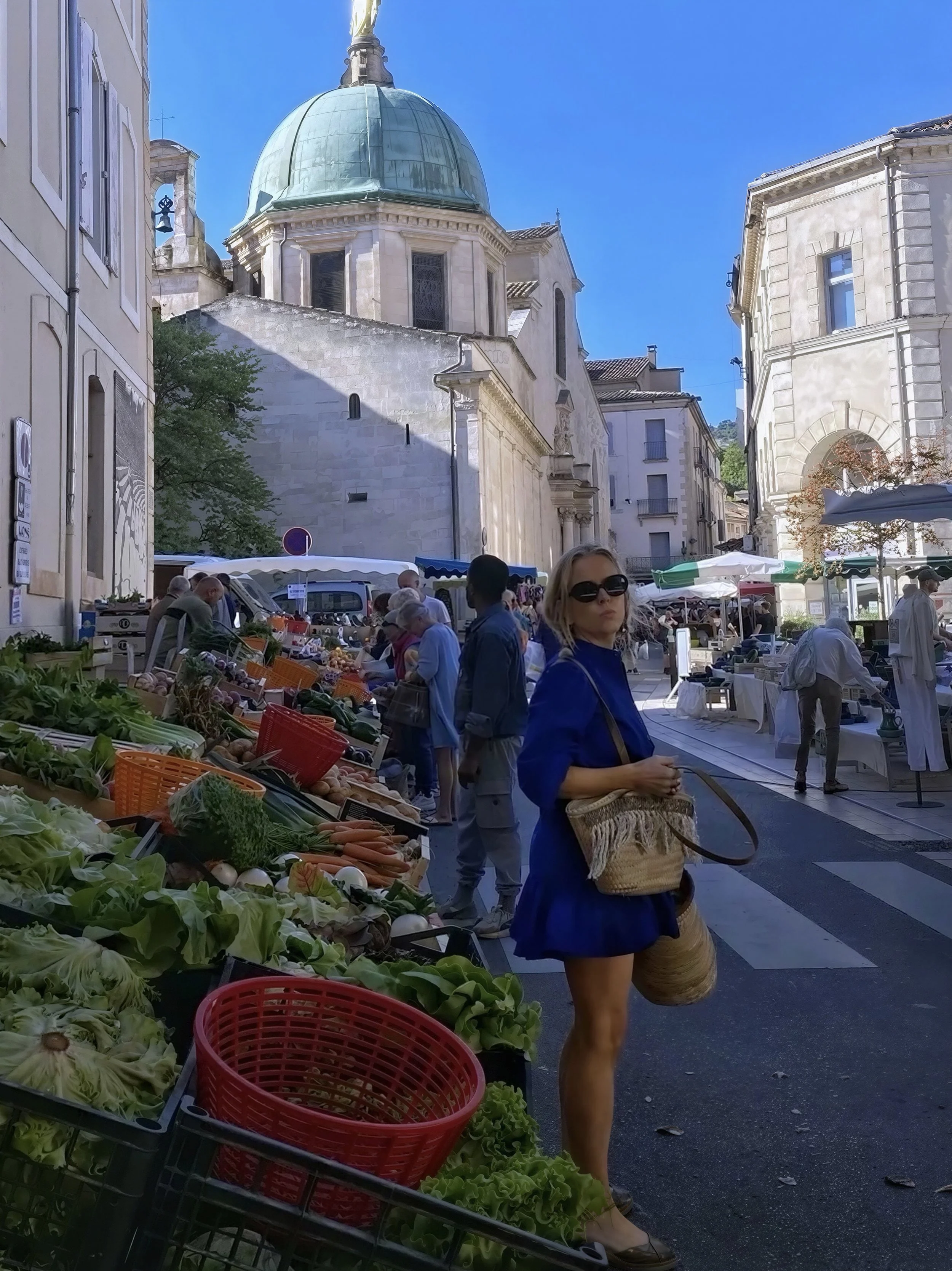 A street market scene in front of historic buildings with a large domed church in the background, people shopping for vegetables and outdoors.