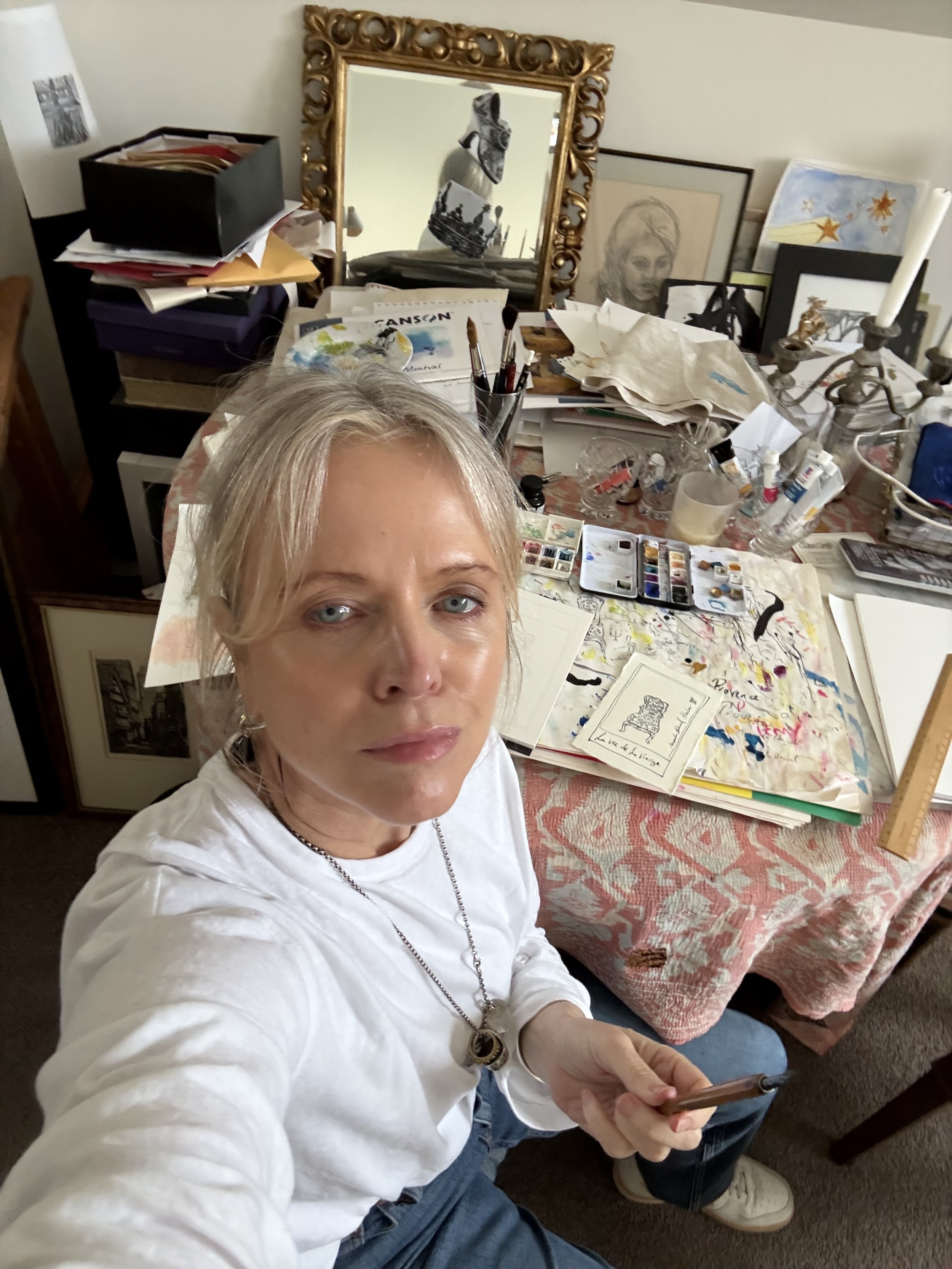 Woman in an artist's cluttered room with a table full of art supplies, framed pictures, and a large mirror in the background.