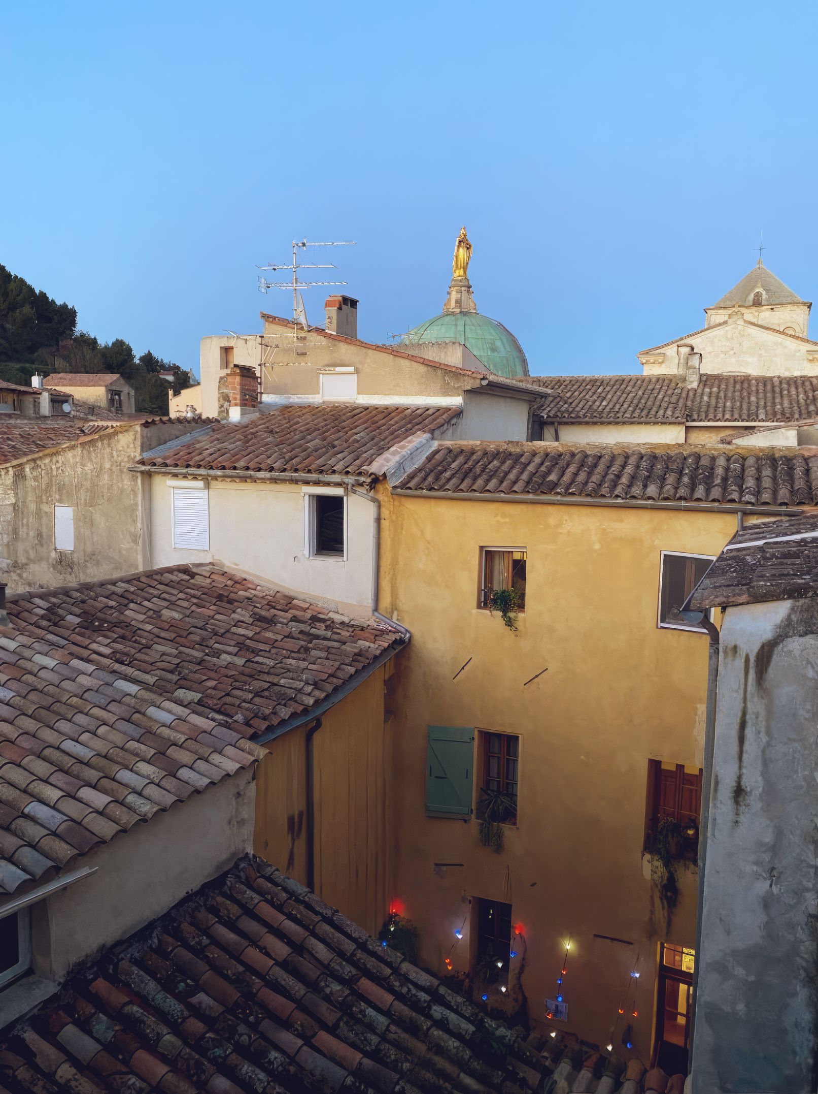View of rooftops in an old Provence European town with tile roofs and a church with a green dome and a golden statue on top, under a clear blue sky.
